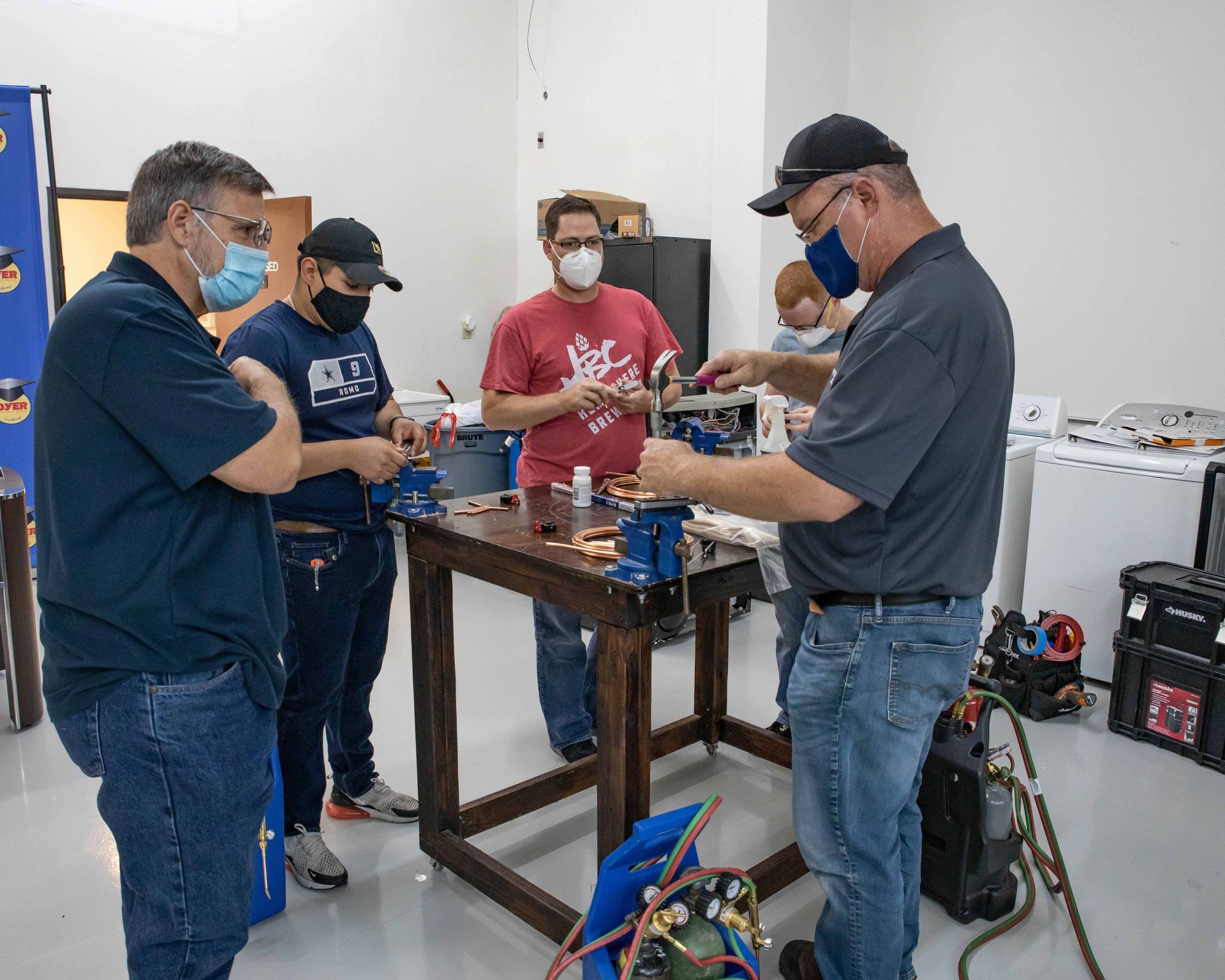 People learning plumbing skills, working at a metal table in a workshop, wearing masks.