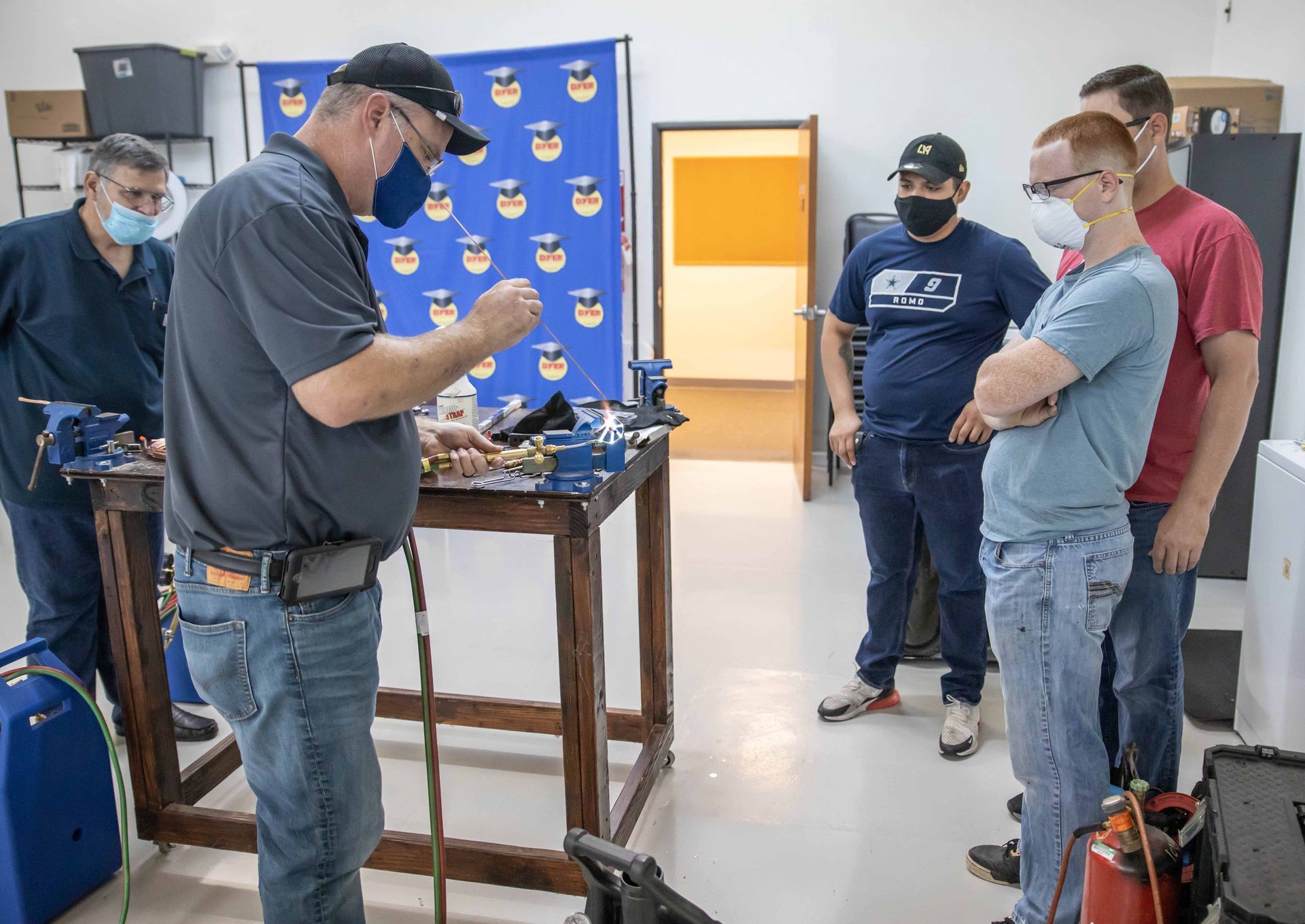 Instructor demonstrating to students at a work table in a workshop, all wearing masks.