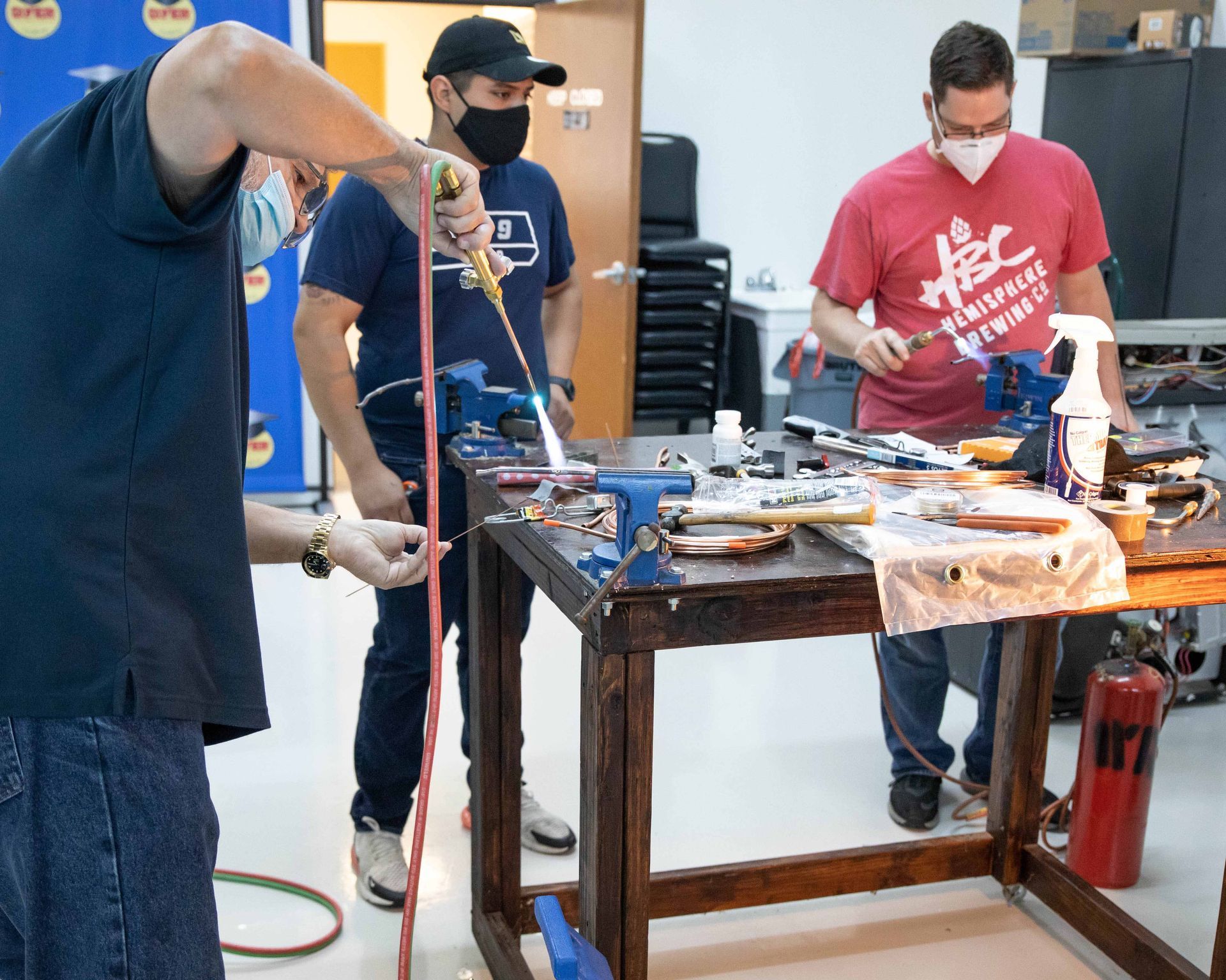 Three people wearing masks working with tools at a workbench. One holds a torch, another watches, and the third works with a piece.