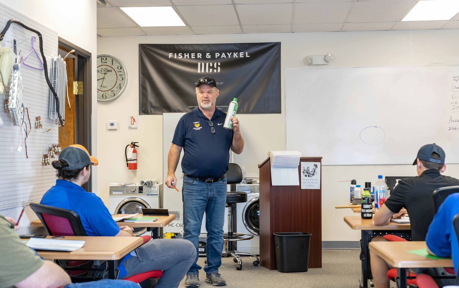 Instructor in a classroom holding a bottle, teaching students. Students seated at desks, taking notes.