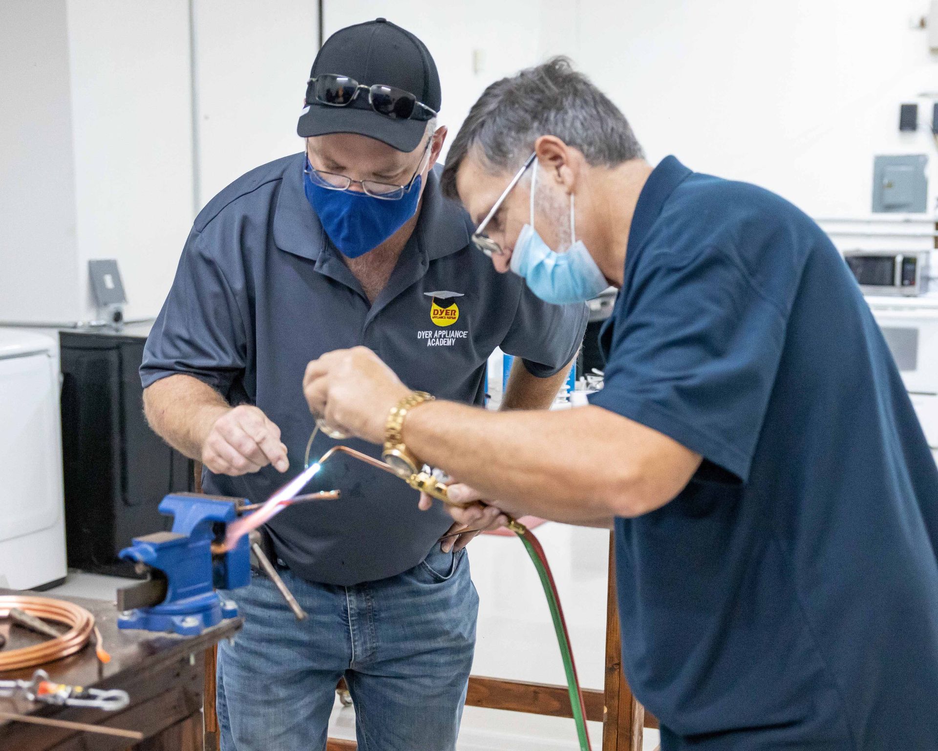 Two people soldering copper pipes in a workshop, both wearing masks and safety glasses.