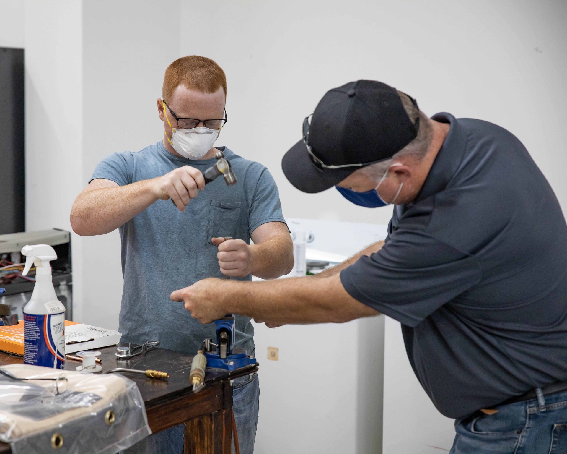 Two men working on a project; one hammering copper, the other points. Indoors, they wear masks and safety glasses.