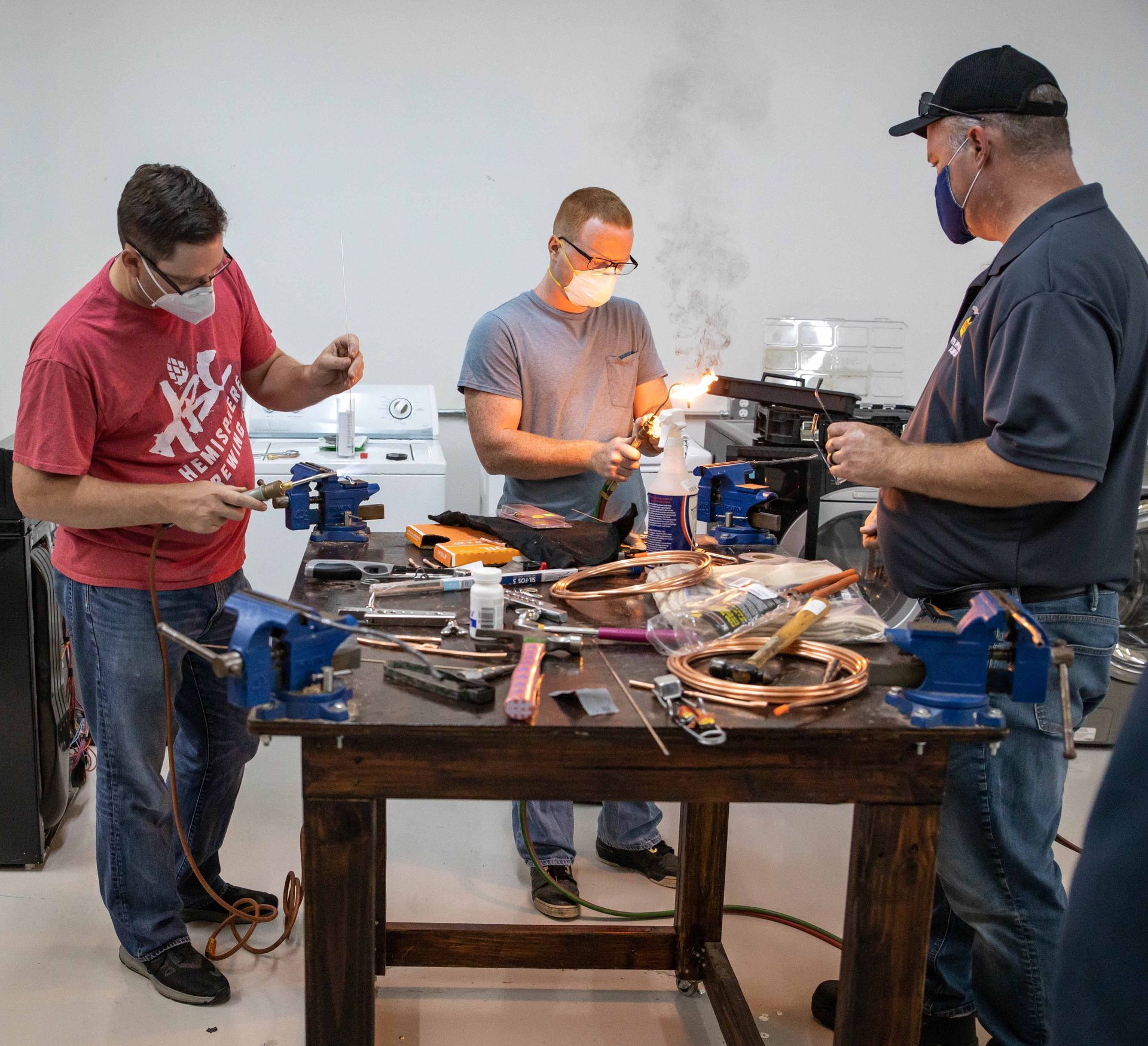 Three people wearing masks, working at a table with tools. One person appears to be soldering.