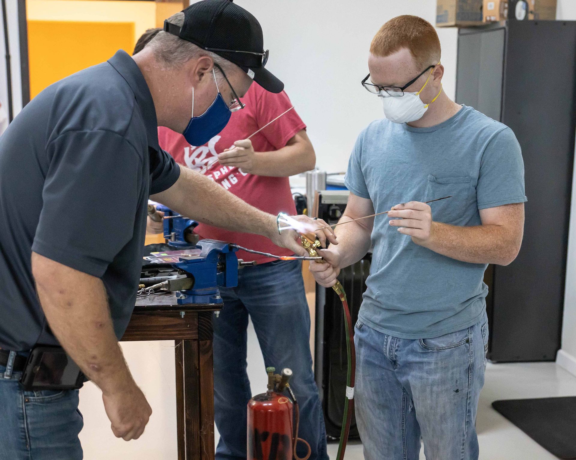 Three people in a workshop setting using tools. One points at a metal rod. Everyone wears masks.