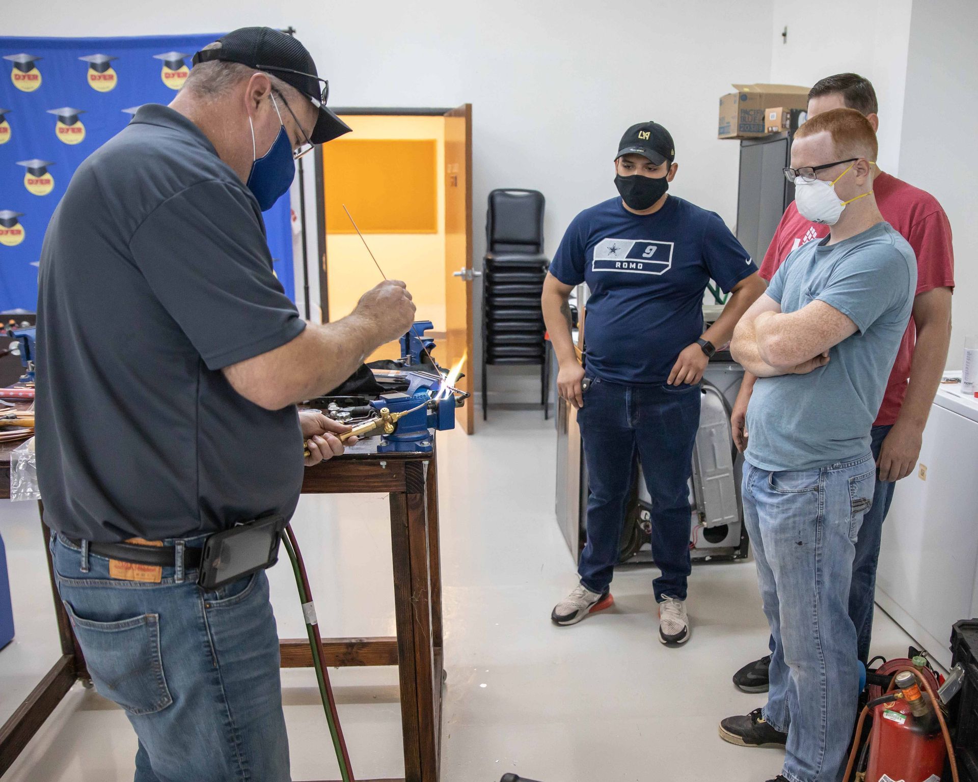 Man demonstrating soldering with three others observing in a workshop setting.
