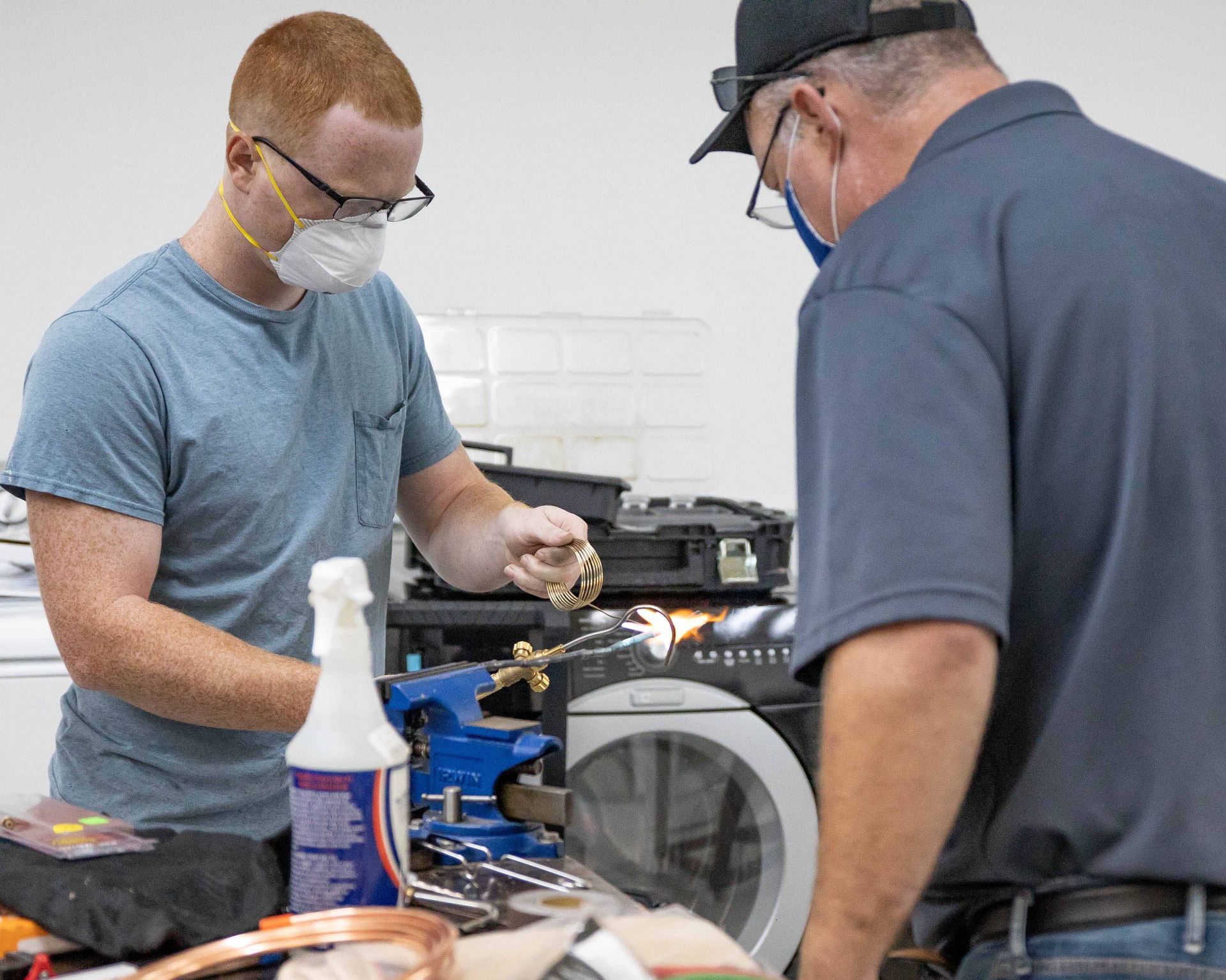 Two men working on equipment; one wearing a mask, glasses, and a blue shirt, the other wearing a hat and shirt, also wearing glasses and a mask.