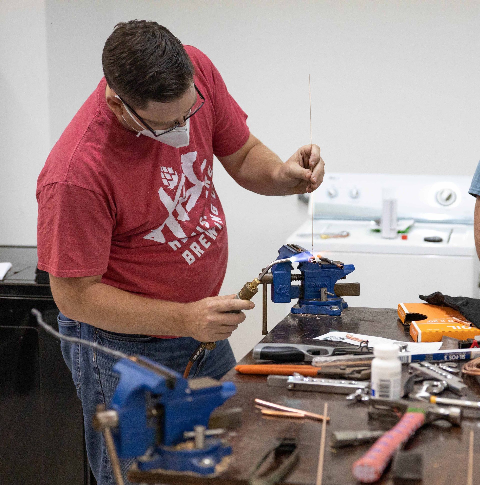 Man soldering copper wire, wearing a mask and red shirt, at a workbench with tools.