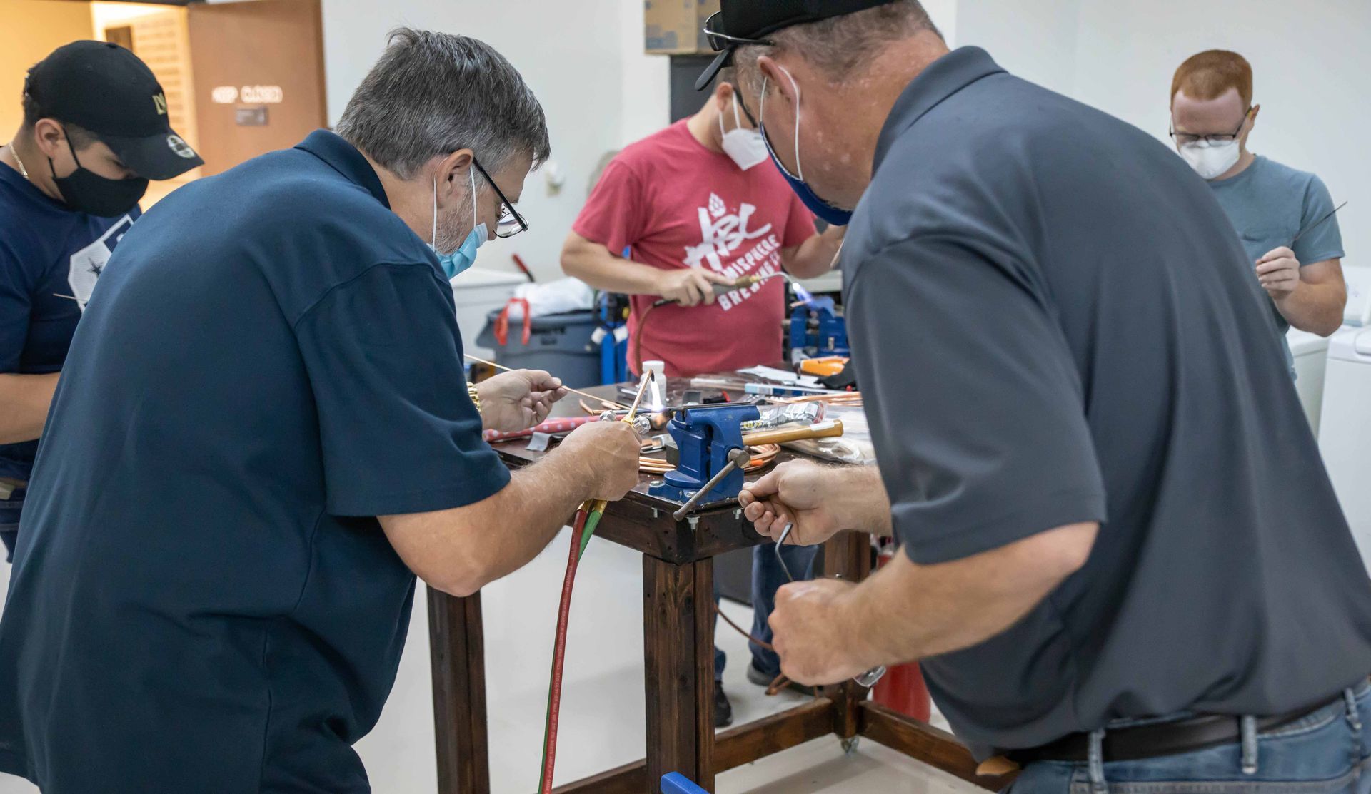 Men working on electrical wiring at a workbench, wearing masks.