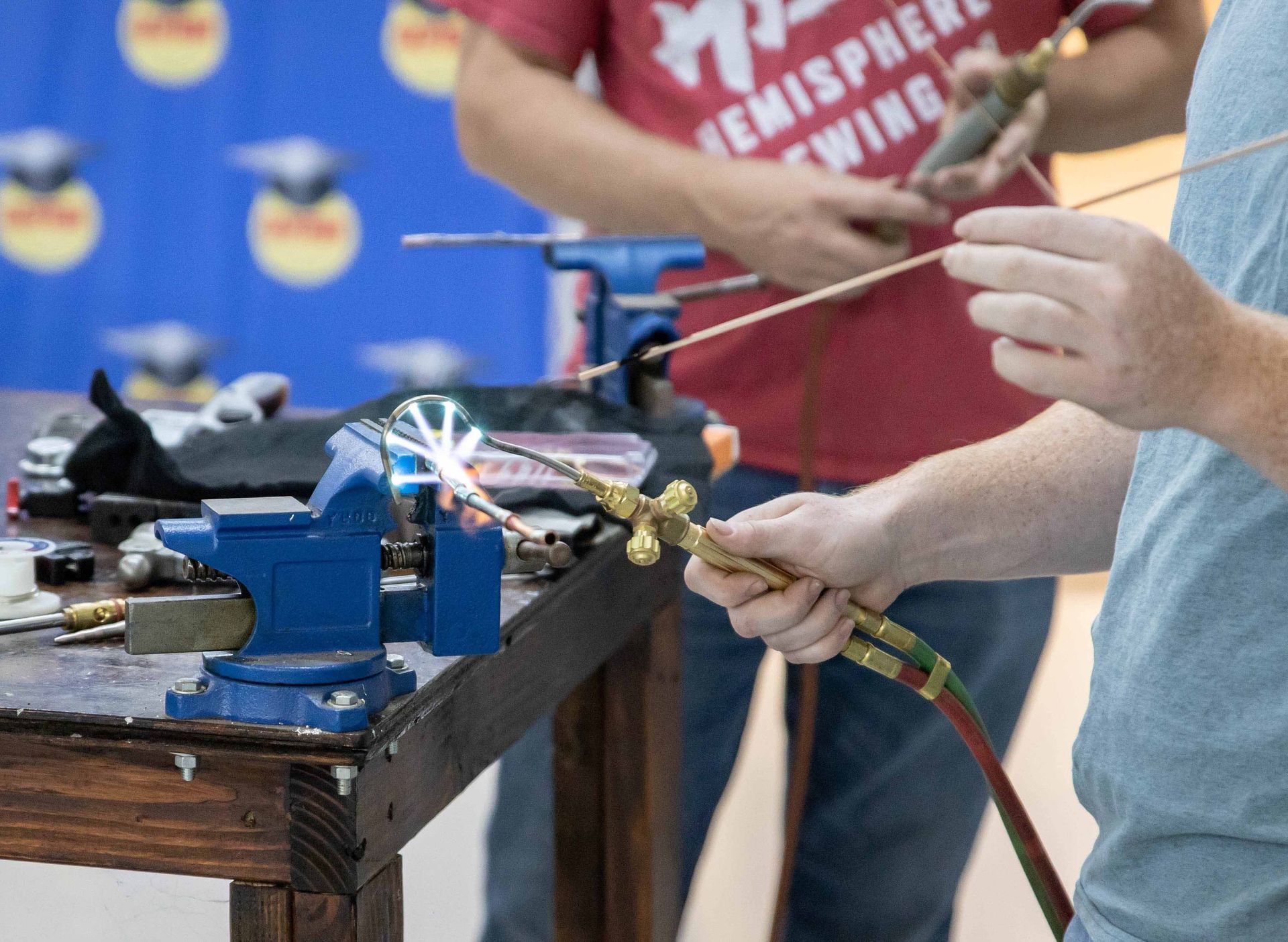 Two people using torches to weld metal clamped in a blue vise.