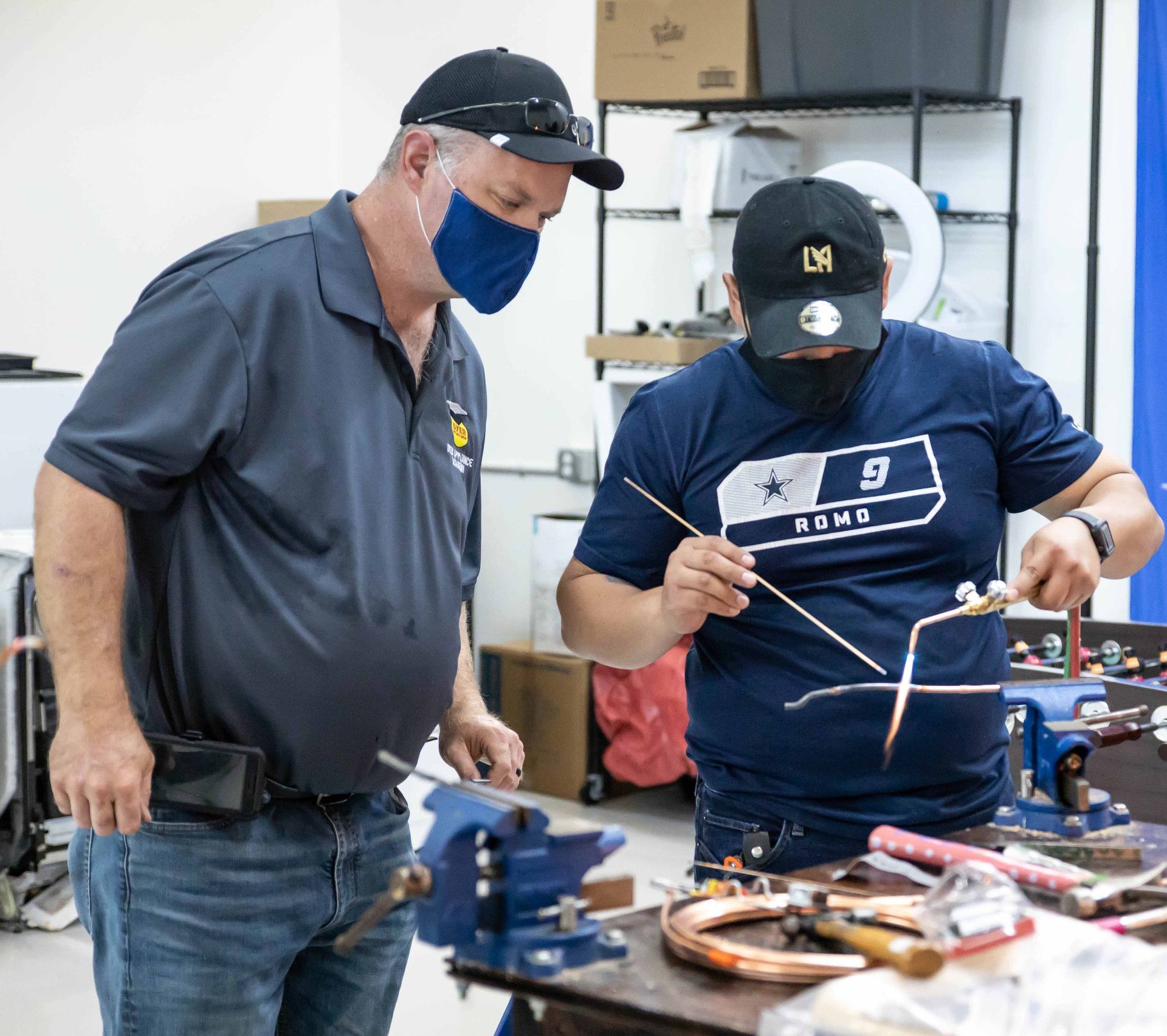 Two men welding copper pipes in a workshop, one observing, both wearing masks and caps.