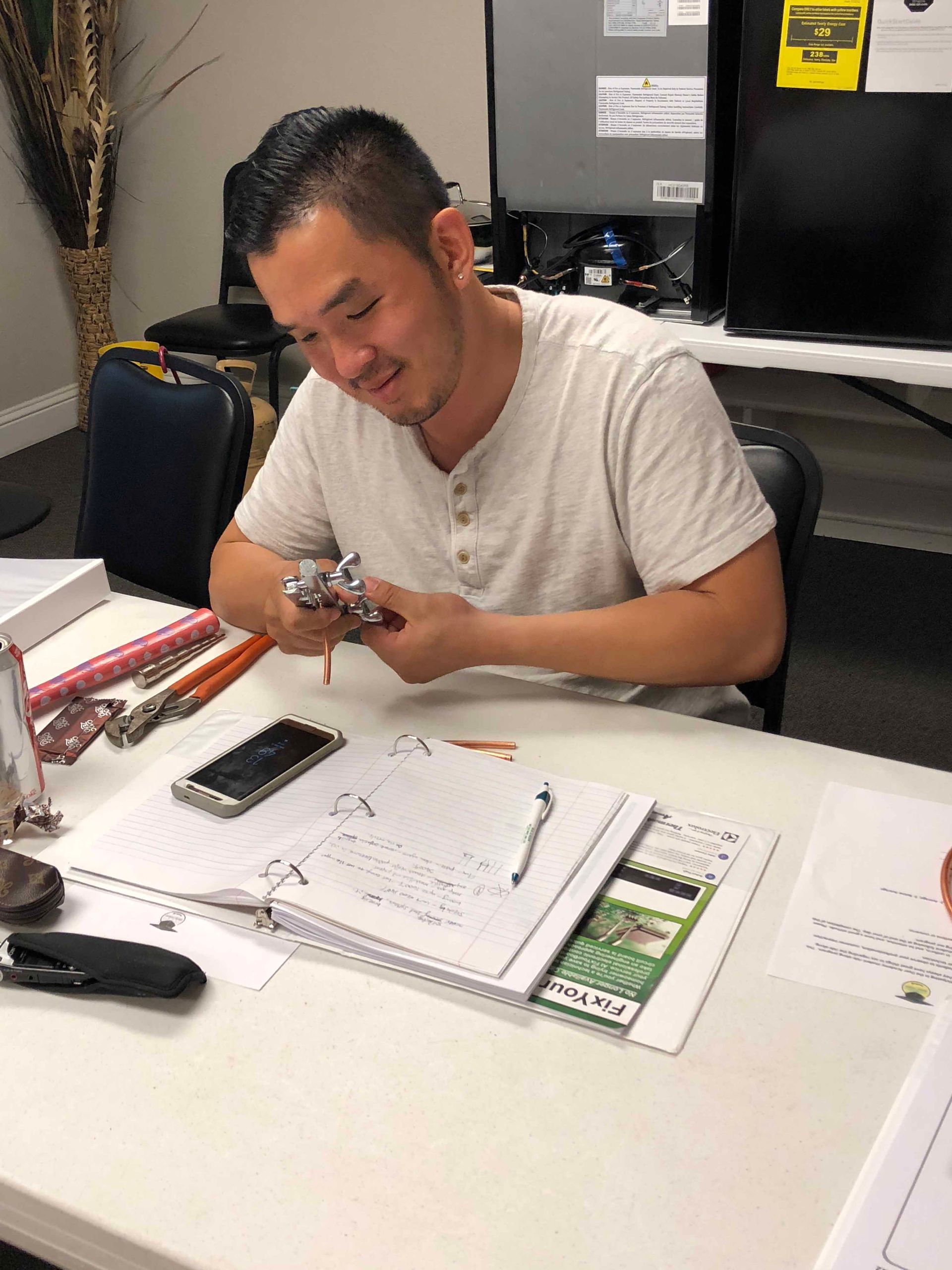 Man seated at a desk, looking down and working on a small object. He wears a light-colored shirt and has a focused expression.