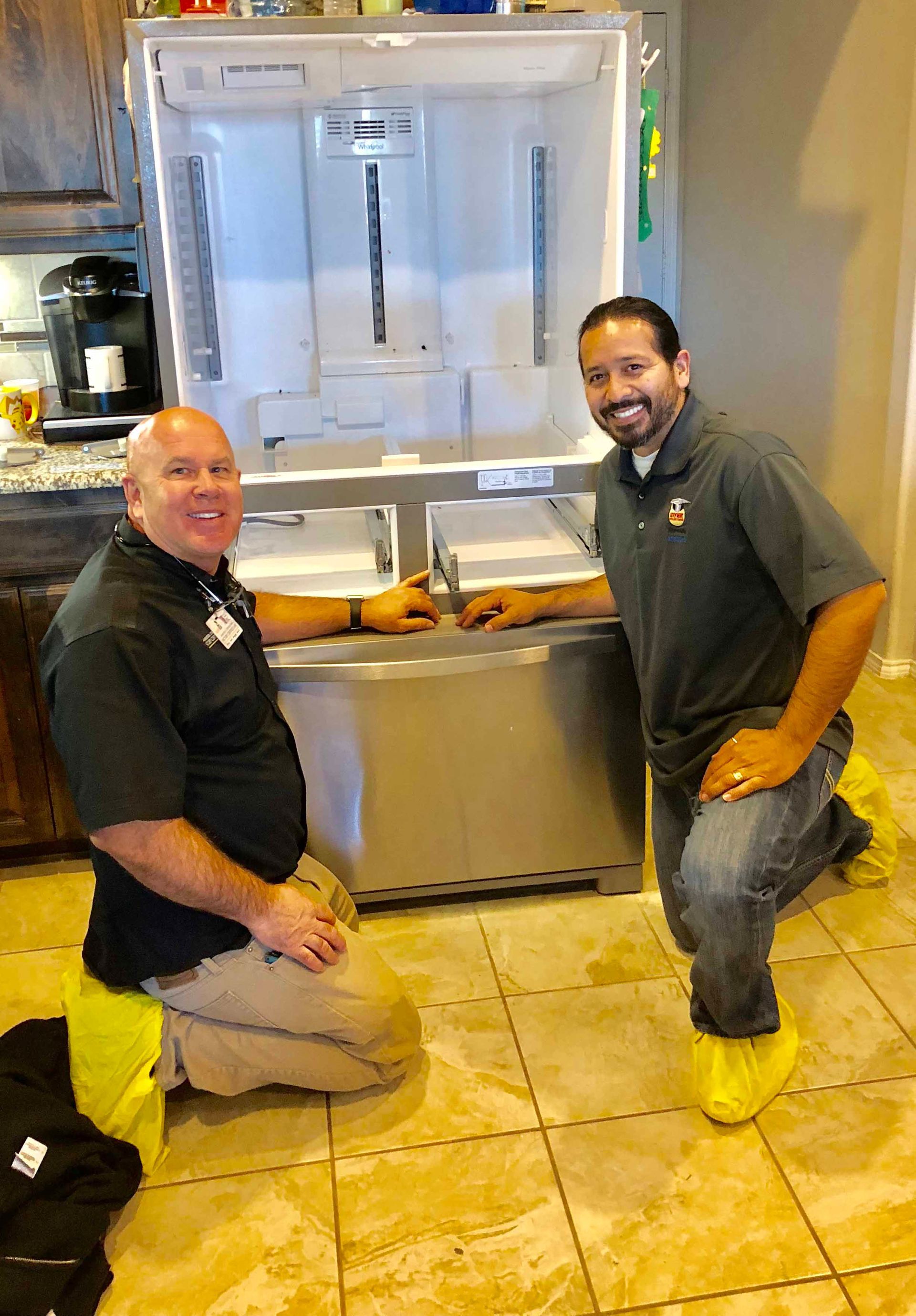 Two men in front of an open refrigerator. They are kneeling, smiling. One wears yellow shoe covers.