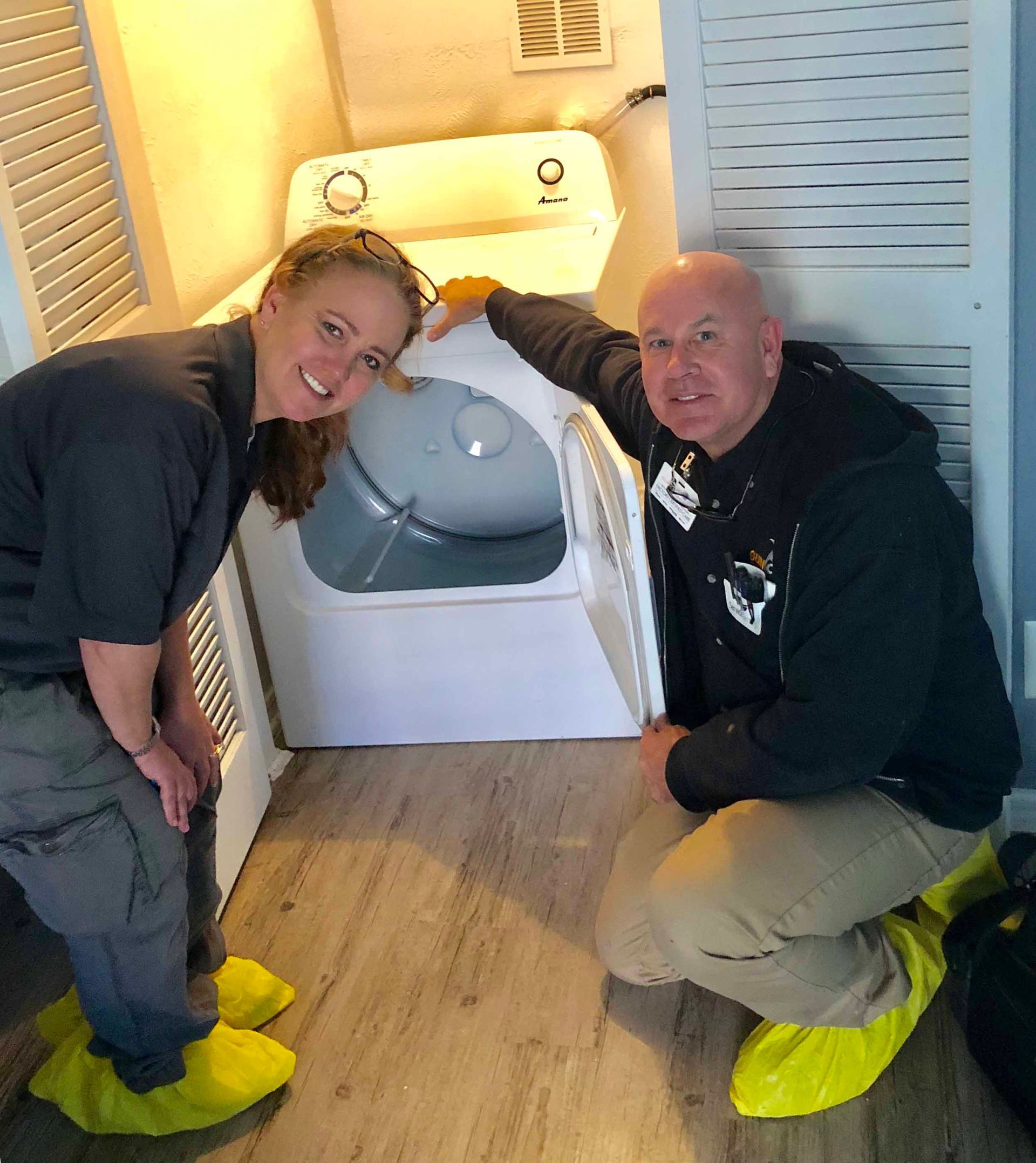Two people kneeling near a dryer, smiling. Both are wearing yellow shoe covers.