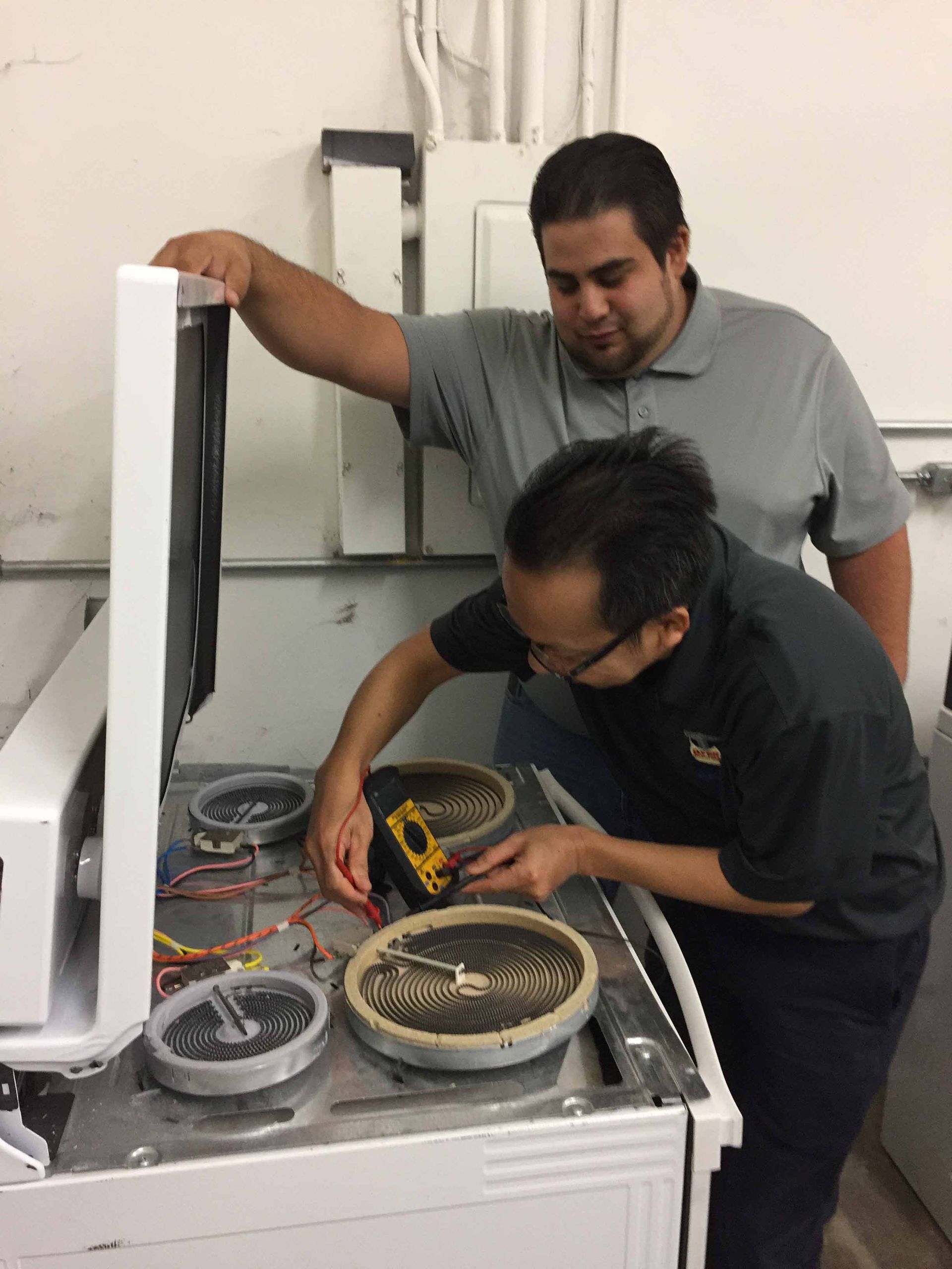 Two people fixing an electric stove. One kneels working on the burners, while the other looks on.