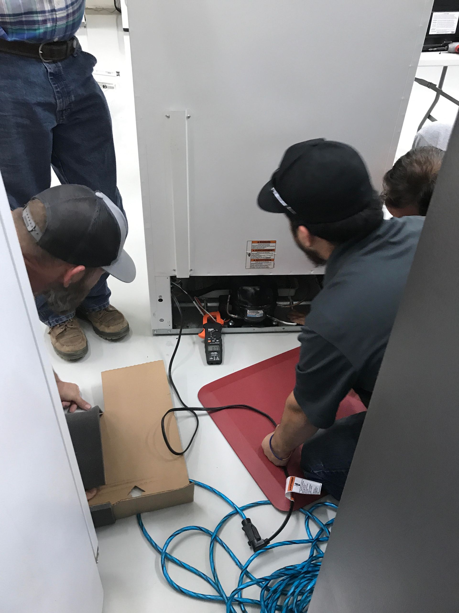 People inspecting the back of a refrigerator, using tools, in a room with white appliances.