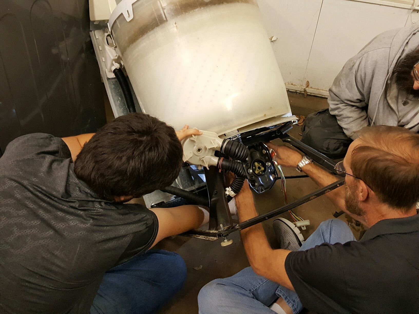 Three people repair a washing machine. They work on the lower mechanism of a white washer in a utility room.