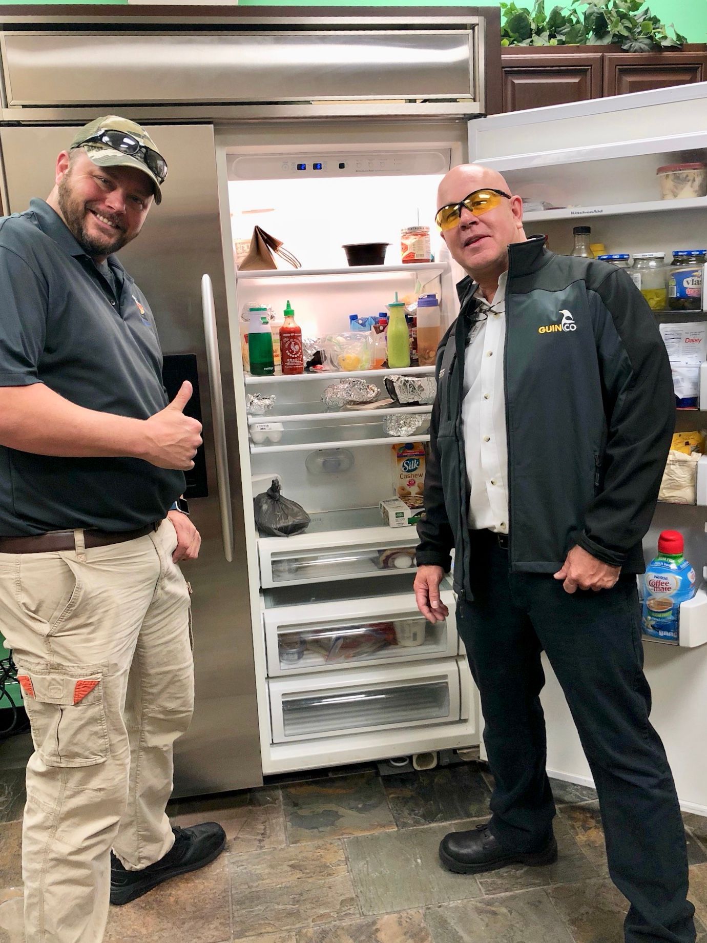 Two men stand by an open refrigerator. One gives a thumbs up. The fridge is stainless steel and stocked with food.