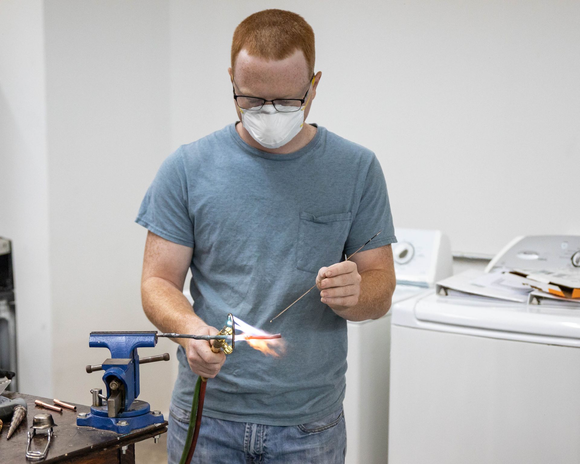 Man wearing a mask and glasses soldering metal in a workshop, with a washing machine visible in the background.