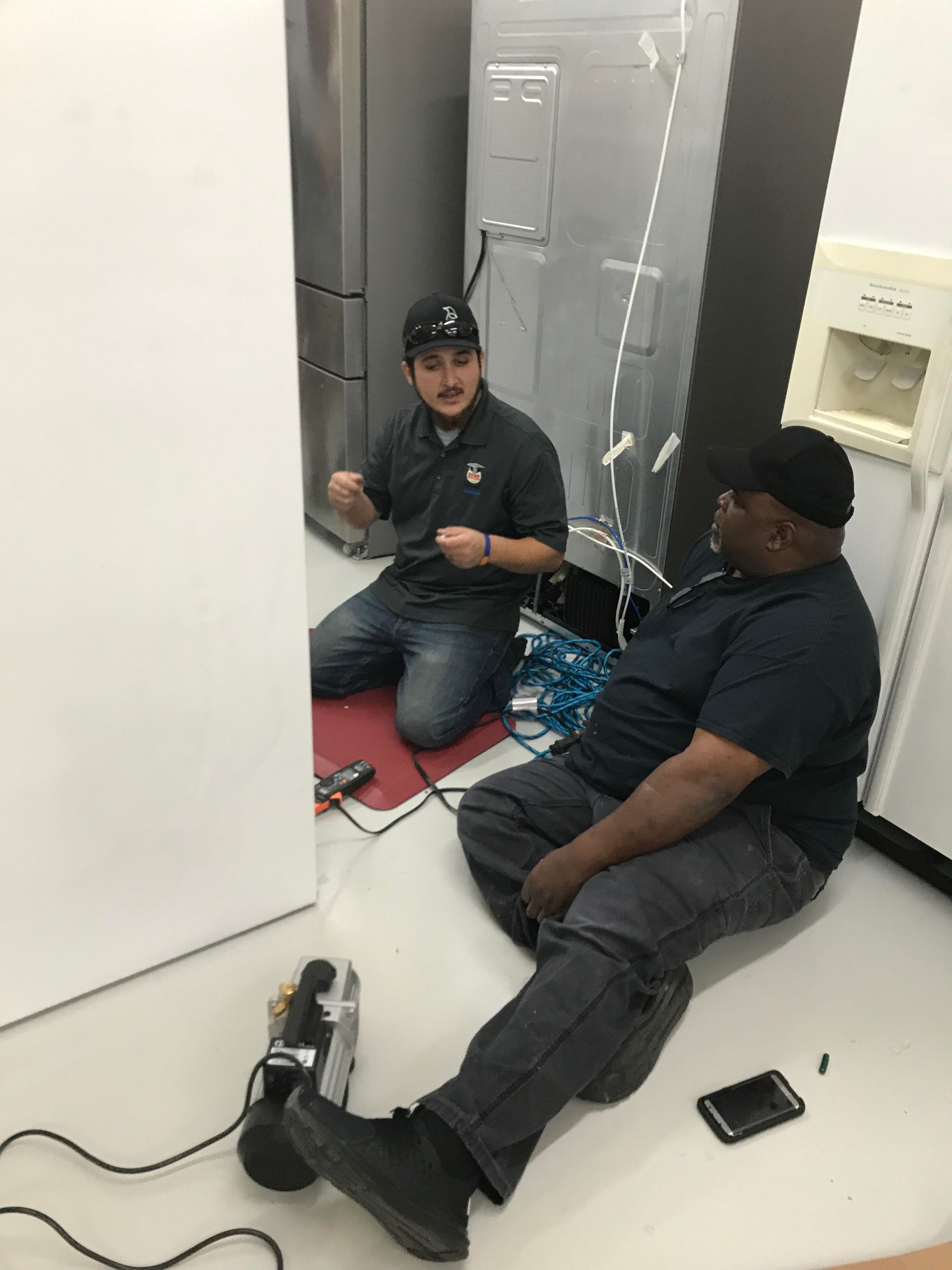 Two appliance repair technicians work on a refrigerator in a white-walled room, one kneeling, one sitting.