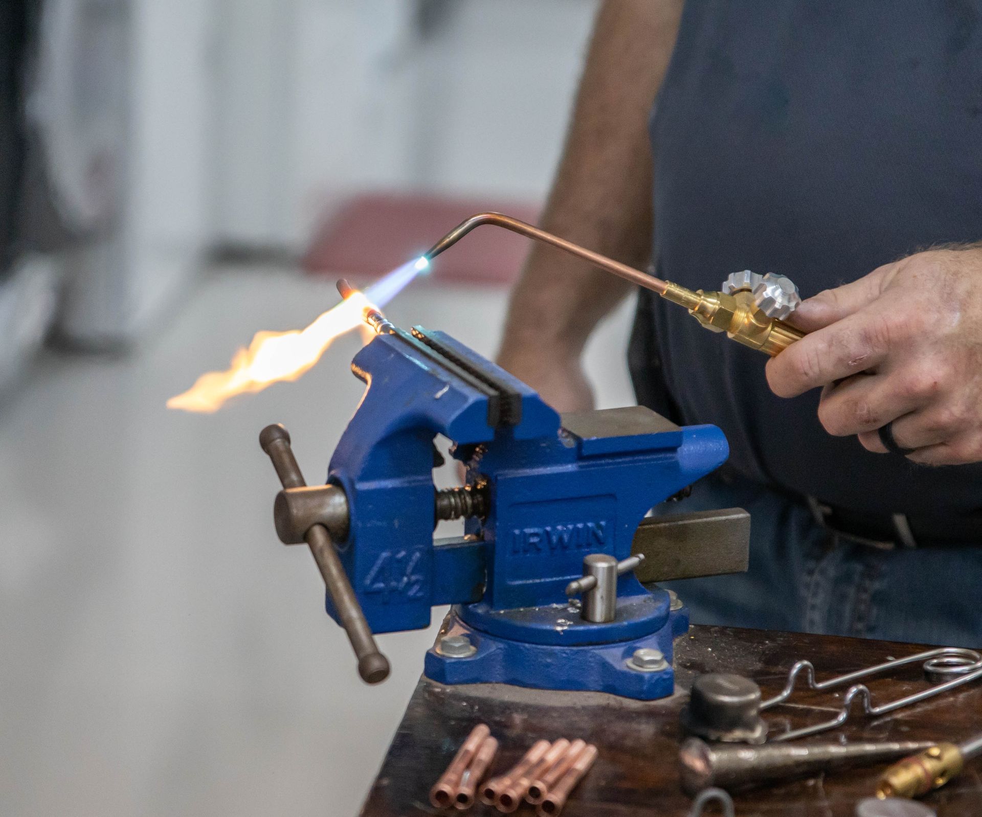 Man using a torch to solder copper pipes clamped in a blue vise on a workbench.