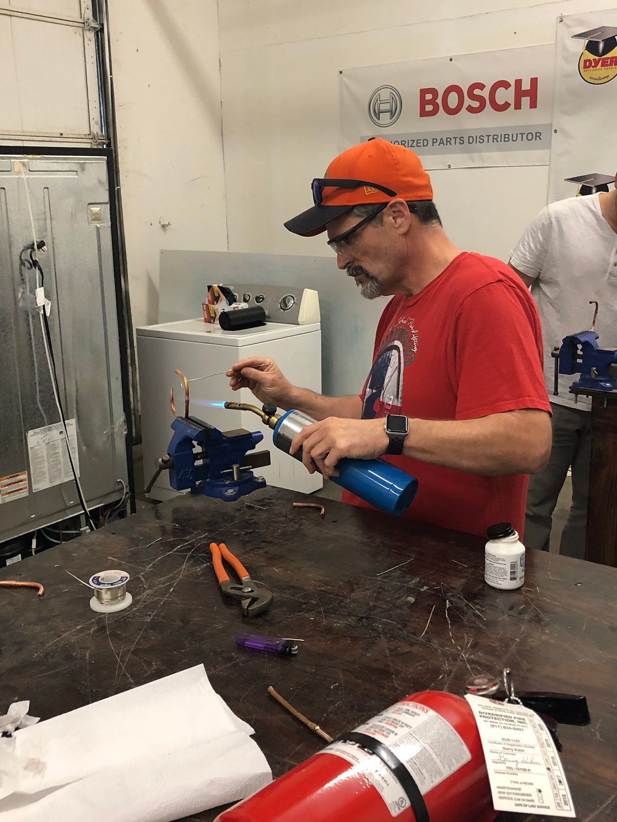 Man soldering copper pipes in a workshop, wearing an orange hat and red shirt. There is a fire extinguisher nearby.