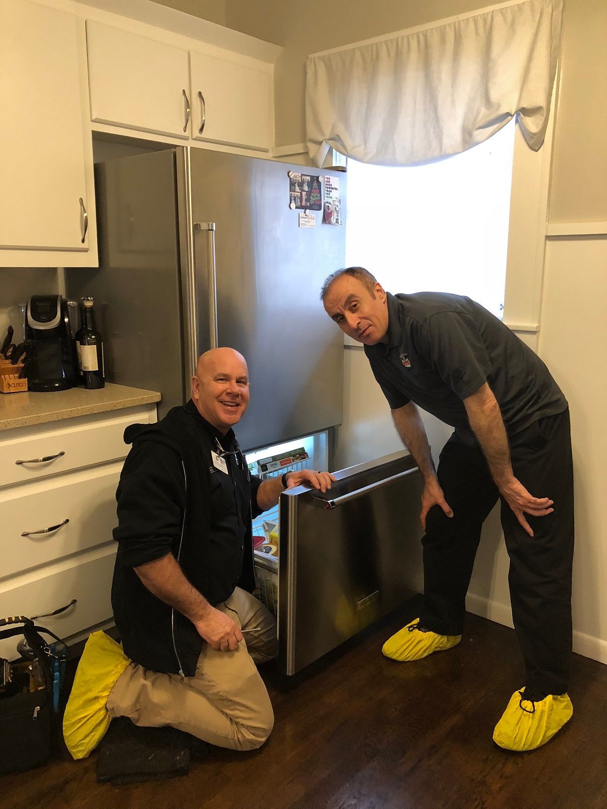 Two men in a kitchen, one kneeling at an open refrigerator drawer. Both are wearing shoe covers.