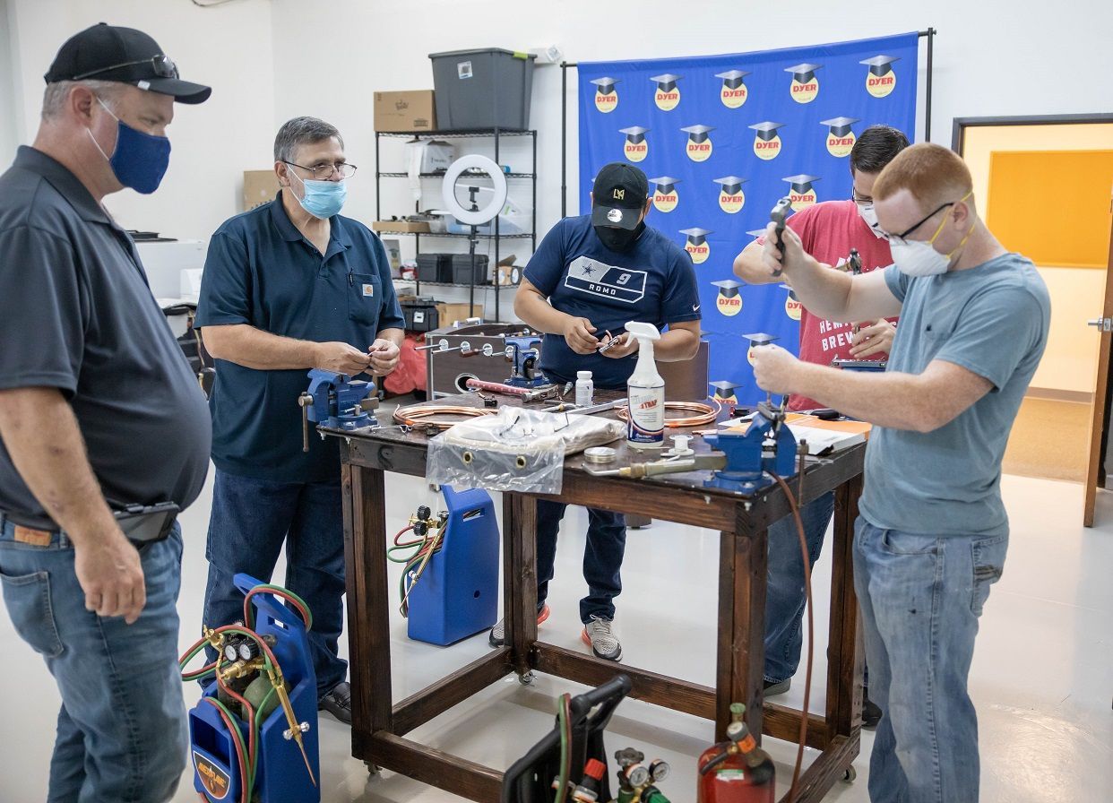 Five people wearing masks work on equipment at a workbench. A blue wall and tools are in the background.