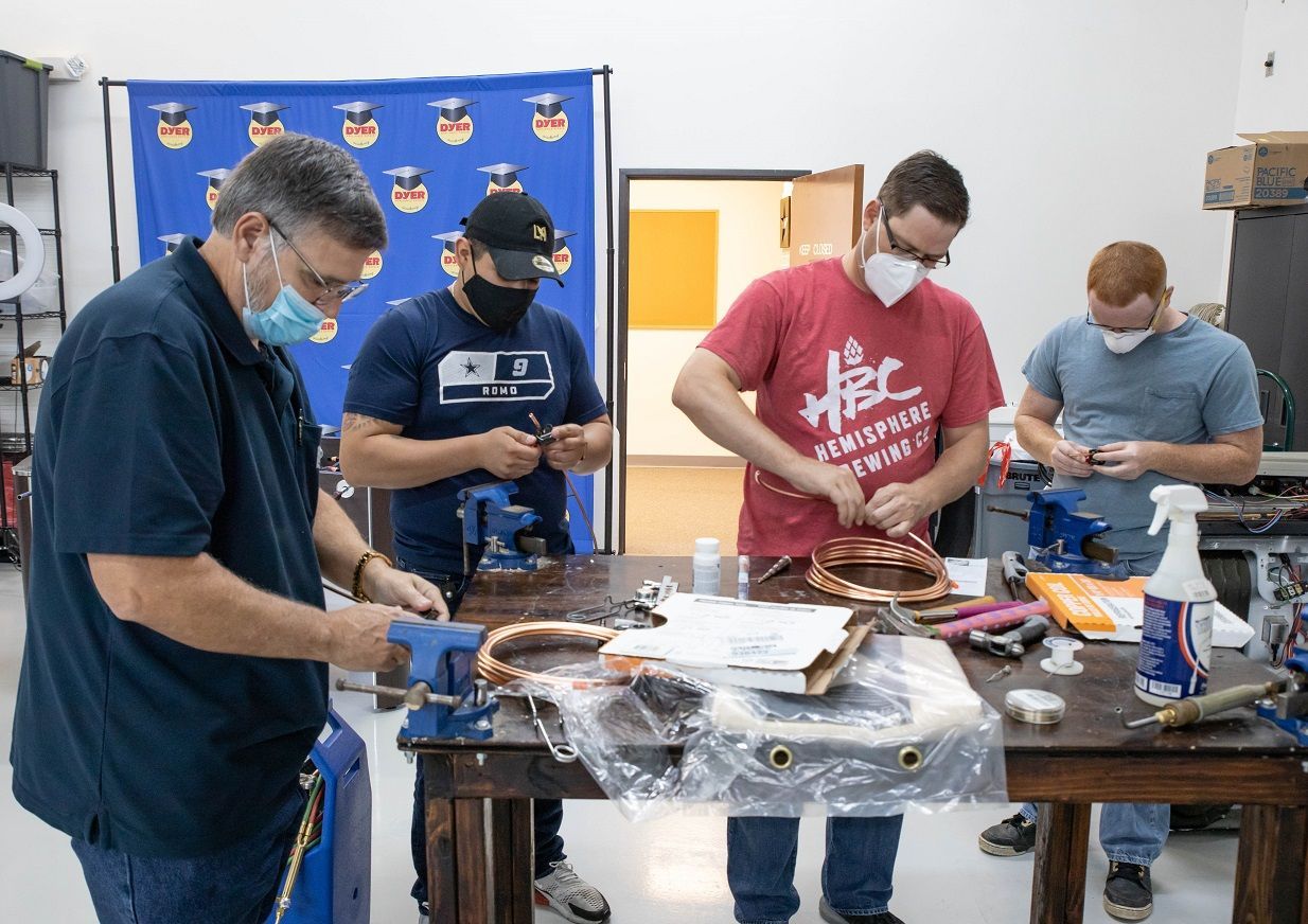Four people work on copper tubing at a workbench, wearing masks and safety glasses.
