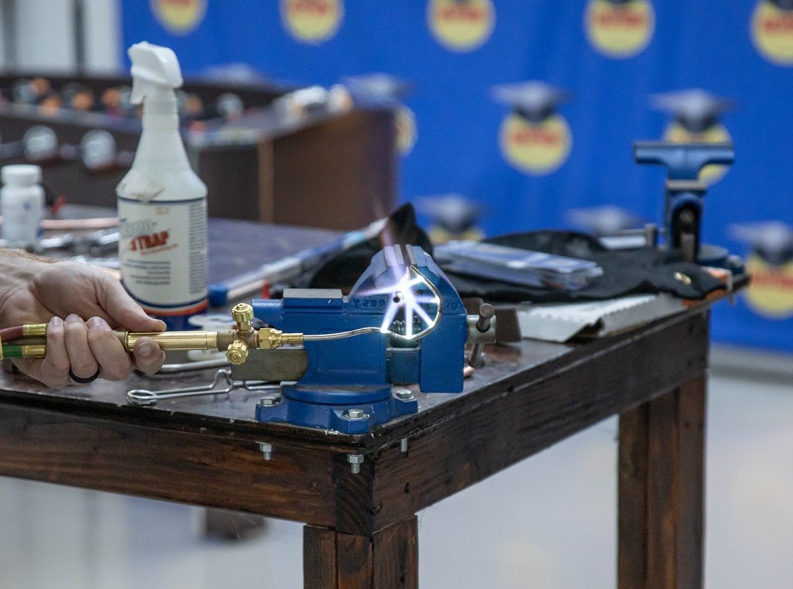 A person uses a torch to heat a metal piece clamped in a blue vise on a wooden table.