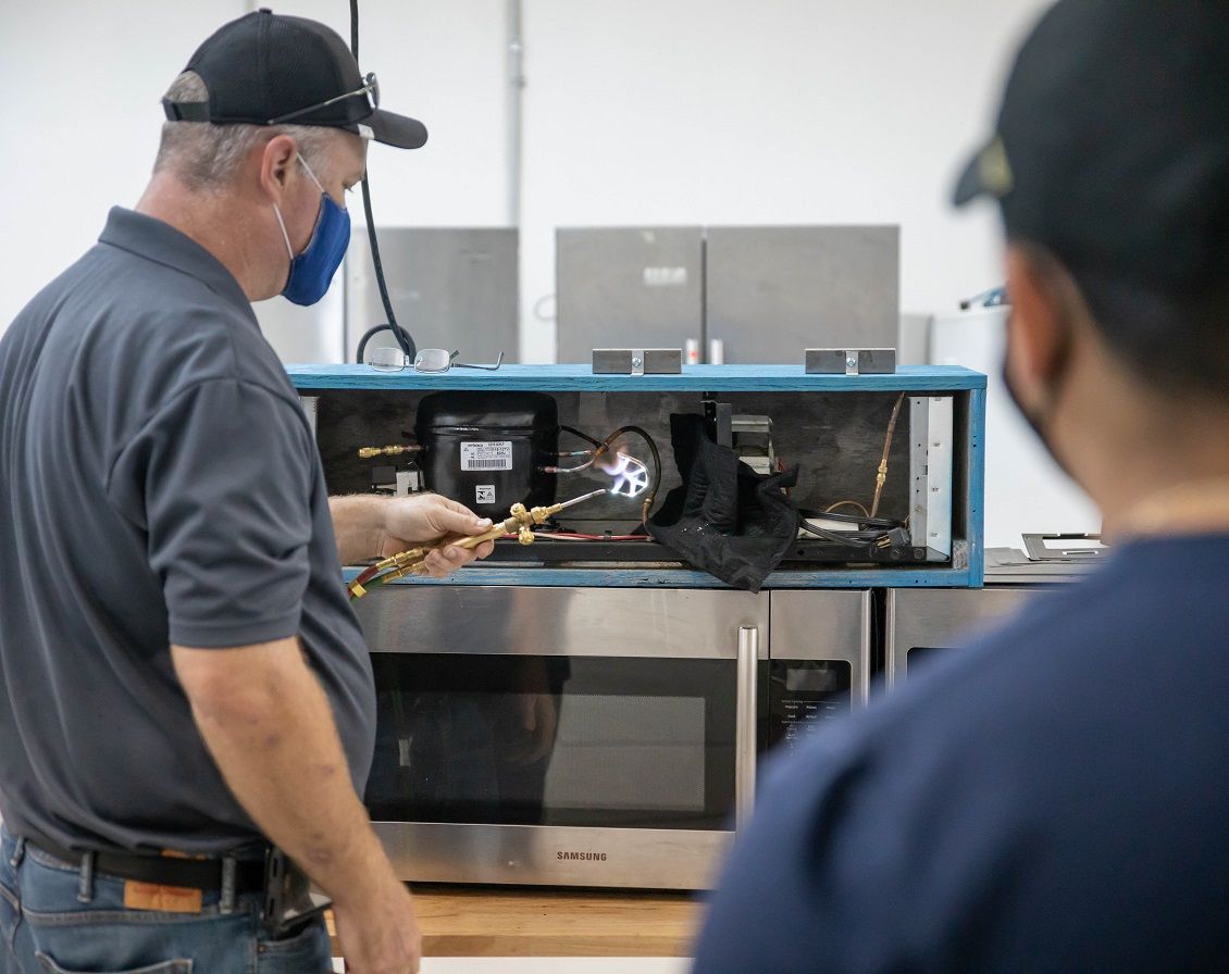 Two people working on refrigeration unit with torch. Indoors, one wearing a mask, another looking on.