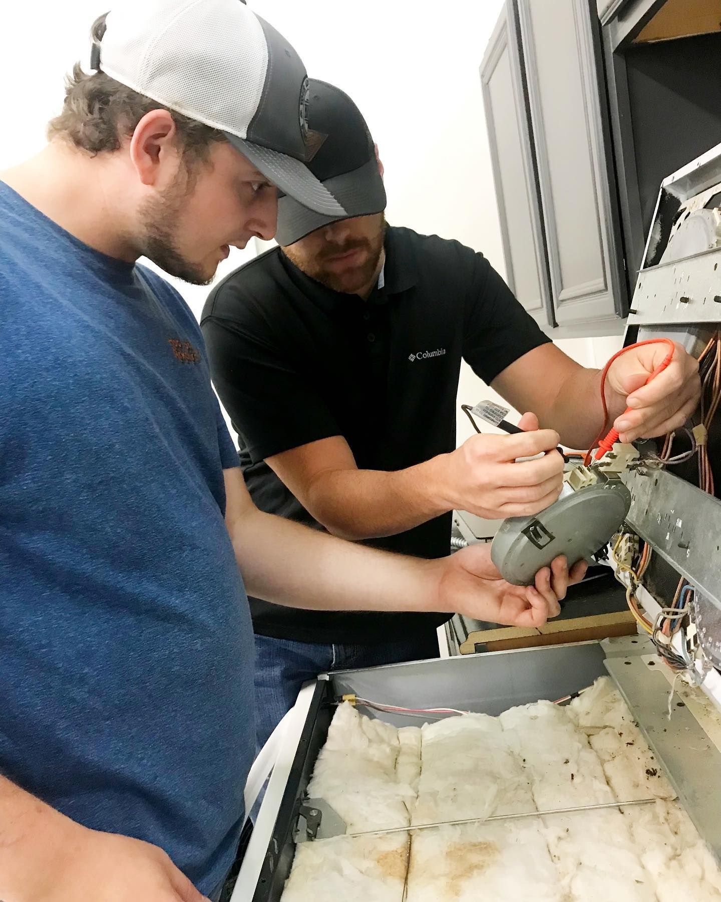 Two men repair an oven, one using a multimeter.