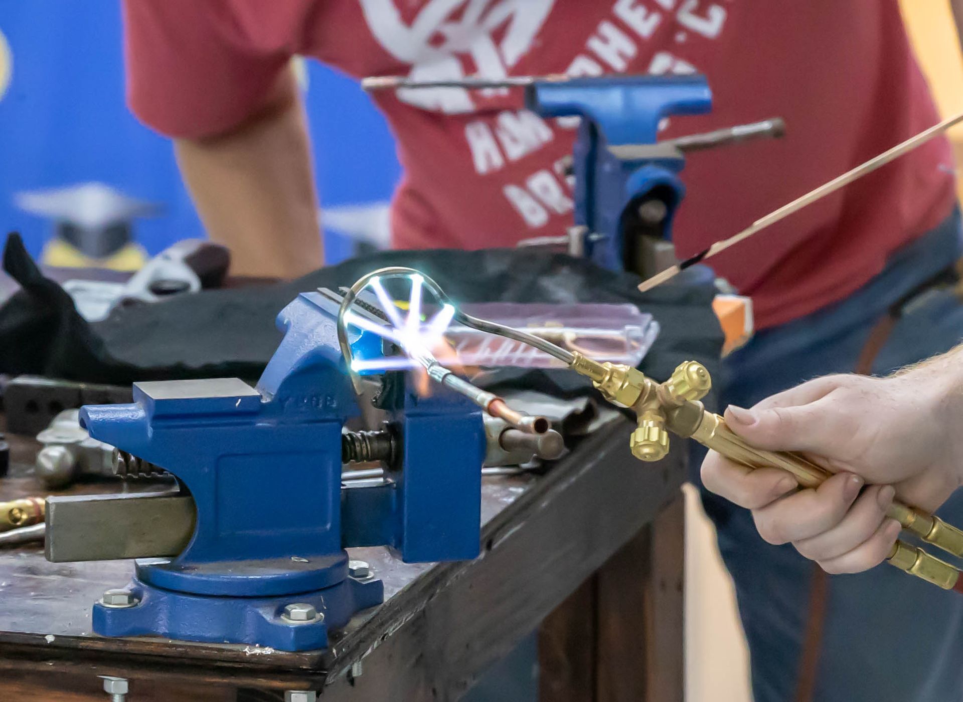 Person using a torch to heat metal clamped in a blue vise on a workbench. Sparks and flames are visible.