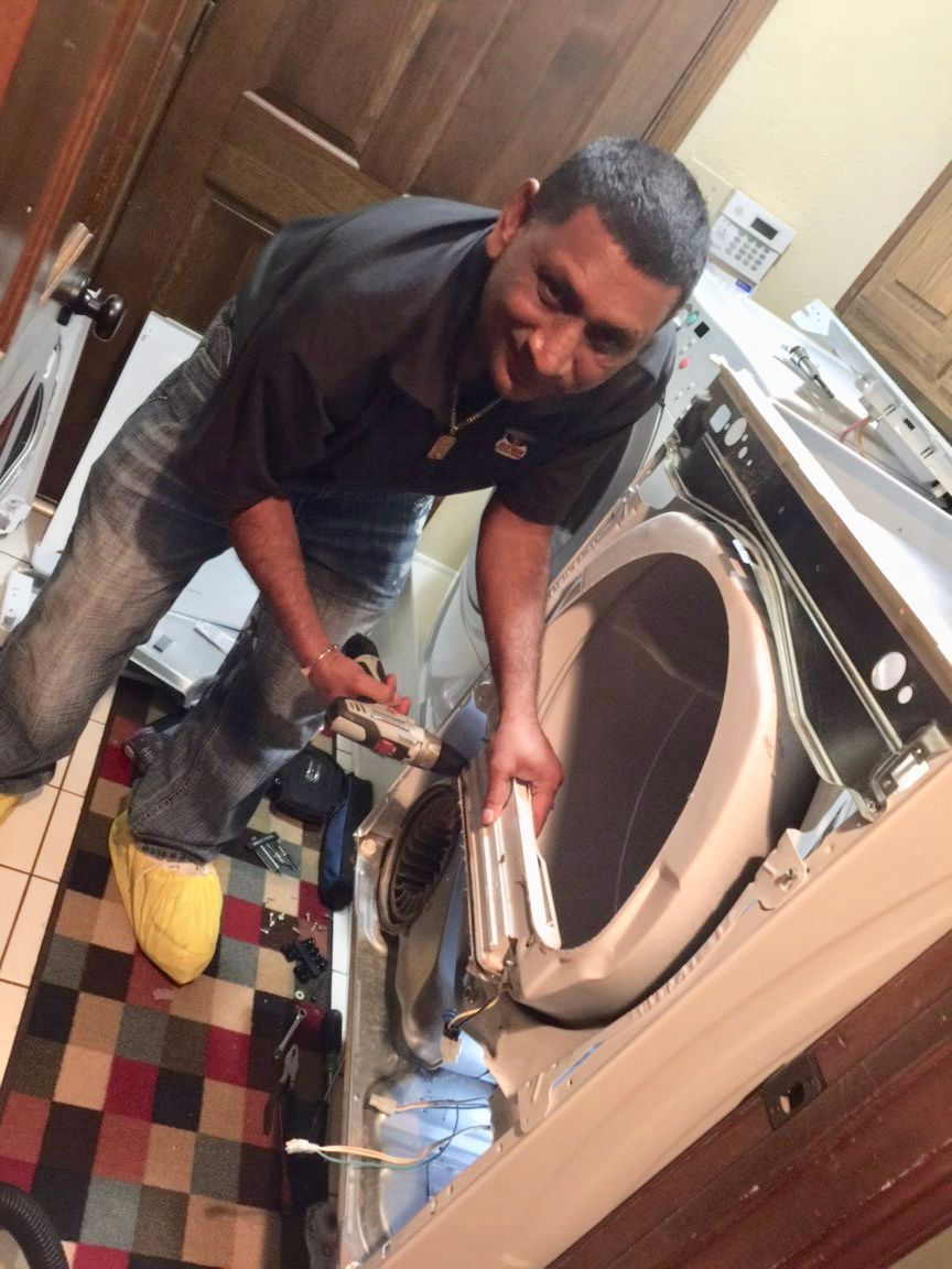 A man fixing a washing machine, smiling. He's inside, with parts spread out, wearing jeans and a black shirt.