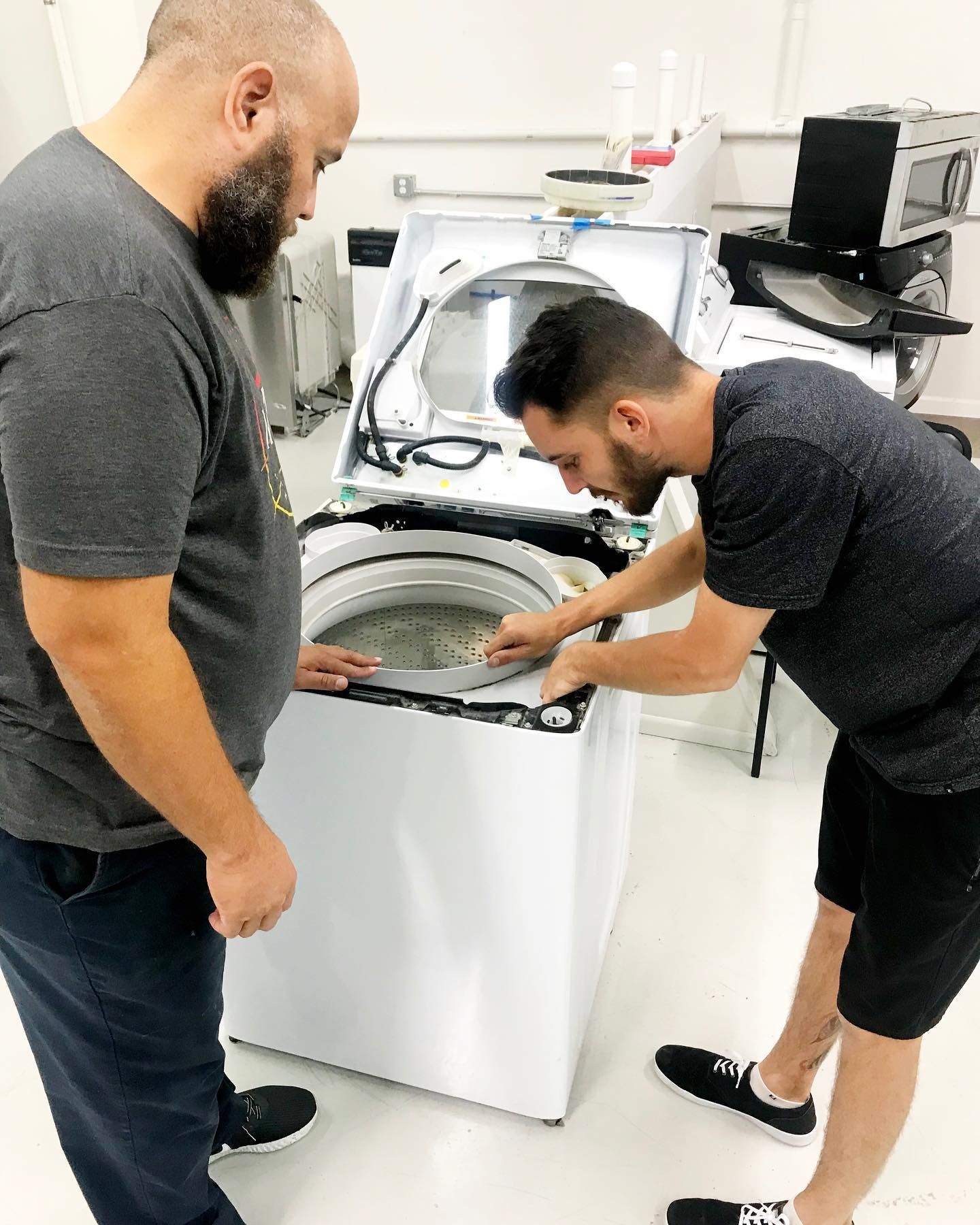 Two men examining the interior of a white washing machine with the lid open in a bright room.