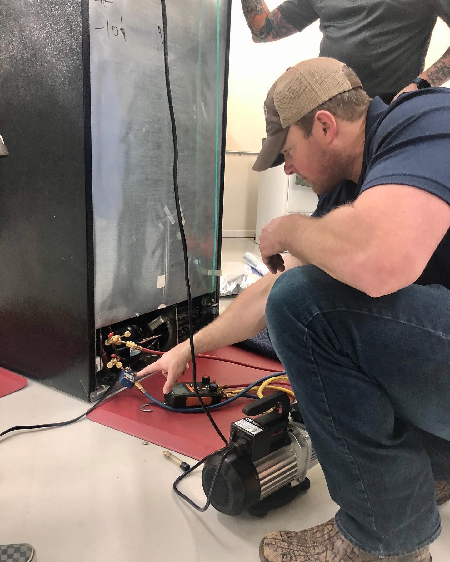 Man in baseball cap points to refrigerator's compressor while another person watches. Vacuum pump is on the floor.