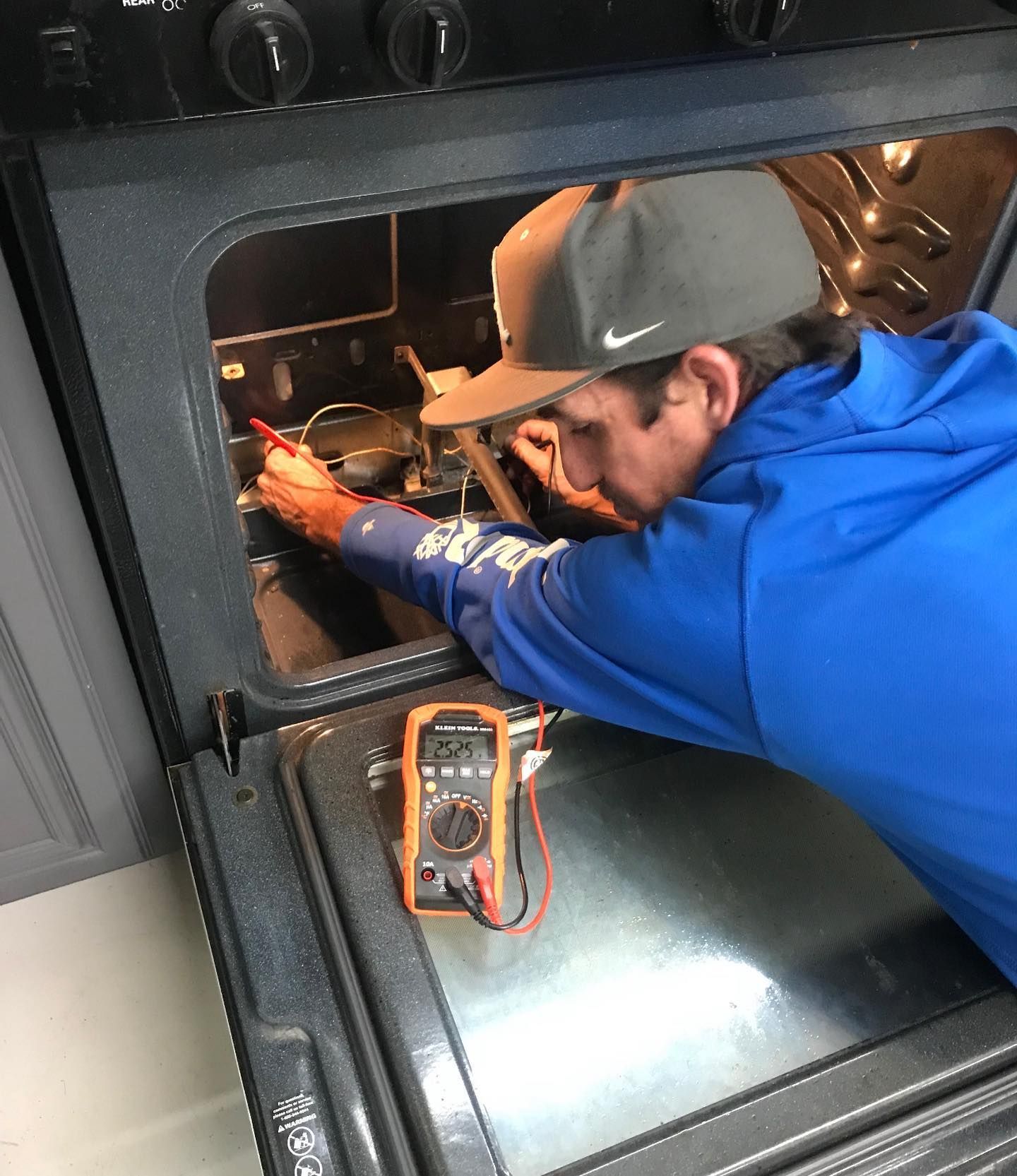 Person in blue shirt and cap uses a multimeter to inspect an oven's interior.