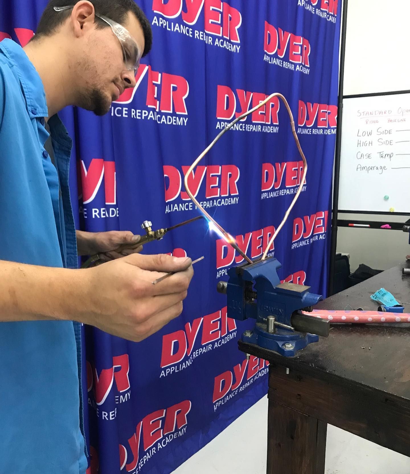 Man in blue shirt using a torch to weld copper tubing clamped in a vise. 