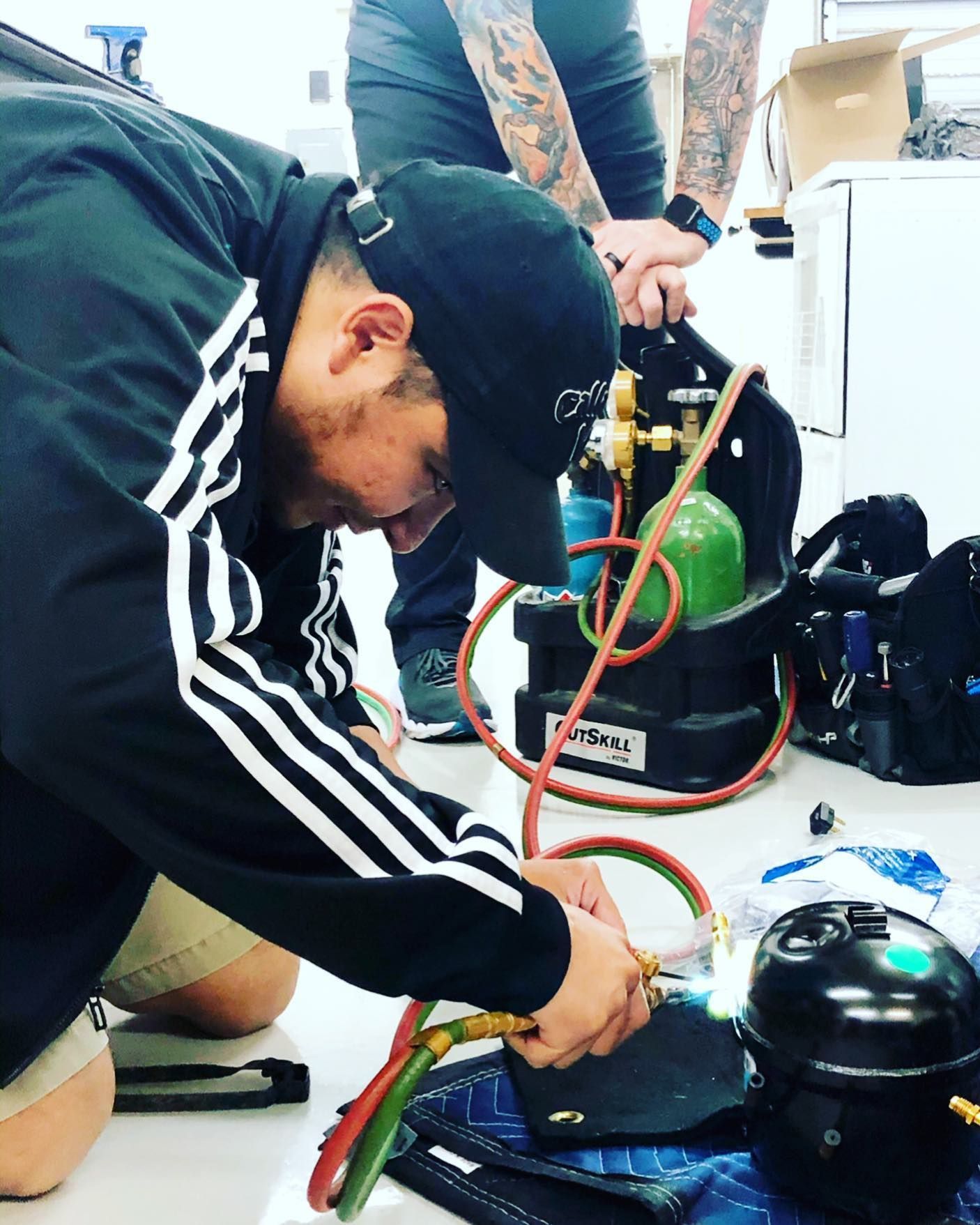 Man in black jacket with white stripes welds with torch. Another person in background.