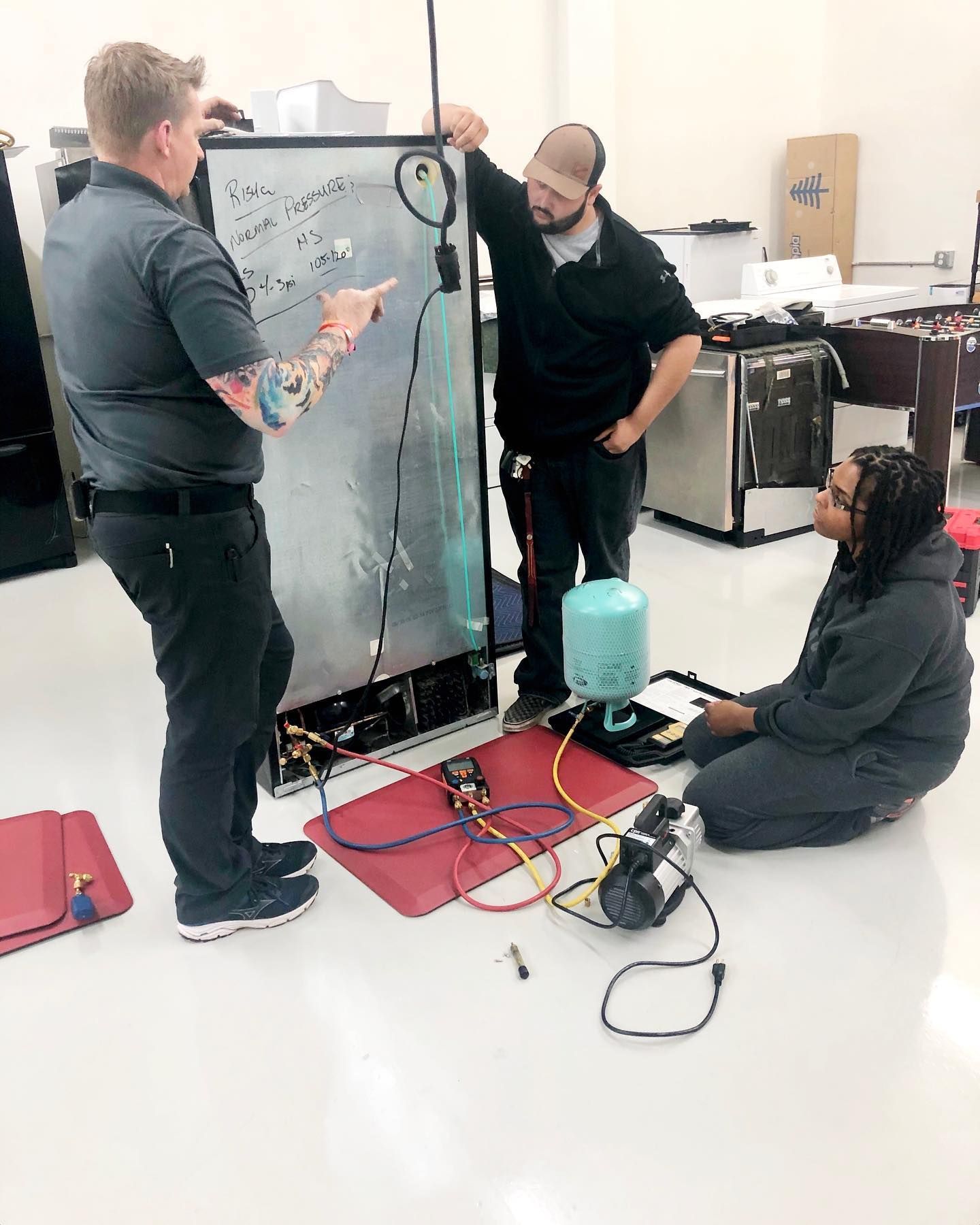 Three people working on appliance repair. A man points at a diagram, while two others observe, one kneeling.