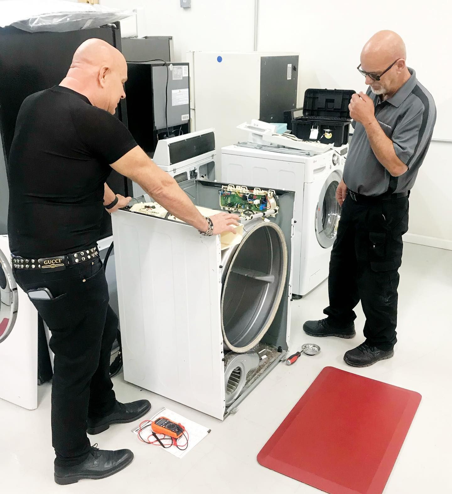 Two men repairing a white washing machine in a workshop, one examining the interior.