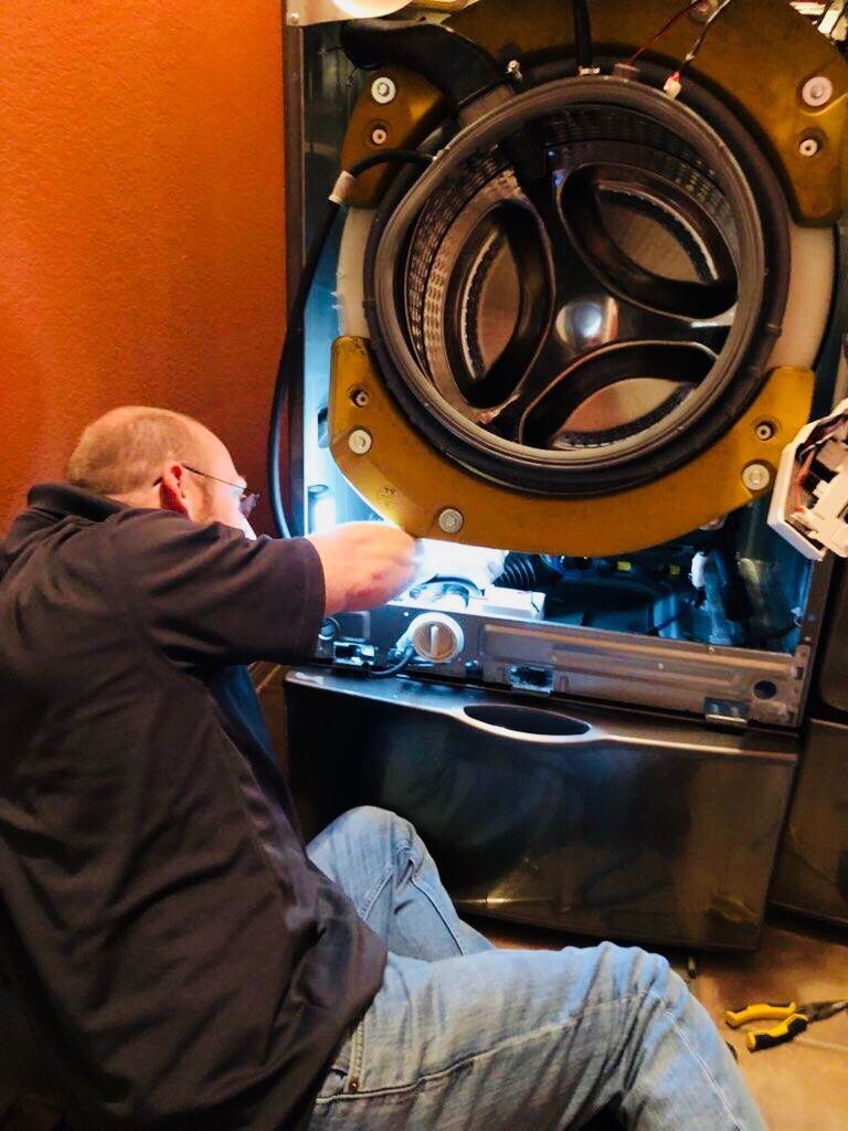 Man repairing washing machine, working inside the appliance. Yellow and black frame visible.