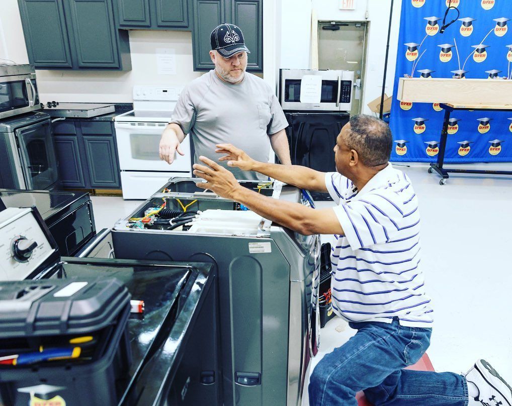 Two men working on appliances in a workshop. One points; the other kneels, gesturing.