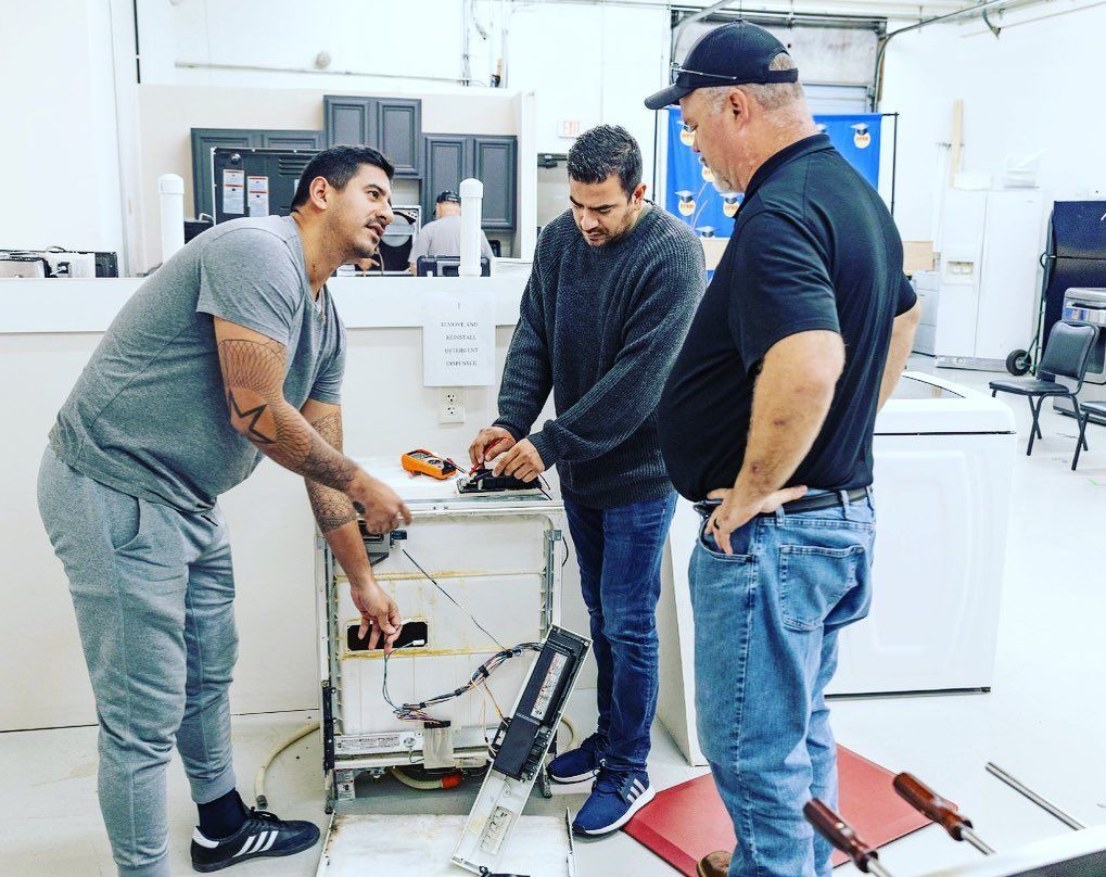 Three people working on appliance wiring in a shop. One points, two examine the unit.