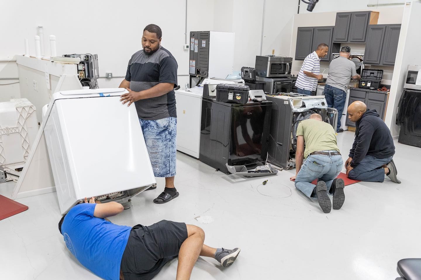 Several men working on appliances in a brightly lit repair shop; one man lifts a washing machine.