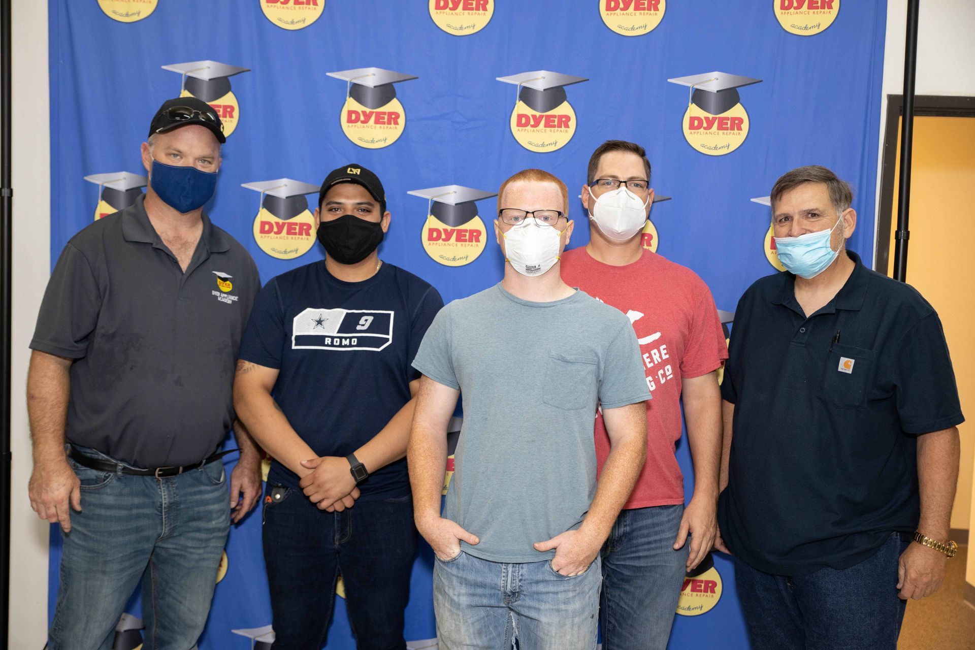 Group of five men wearing masks, standing in front of a blue backdrop with graduation caps.