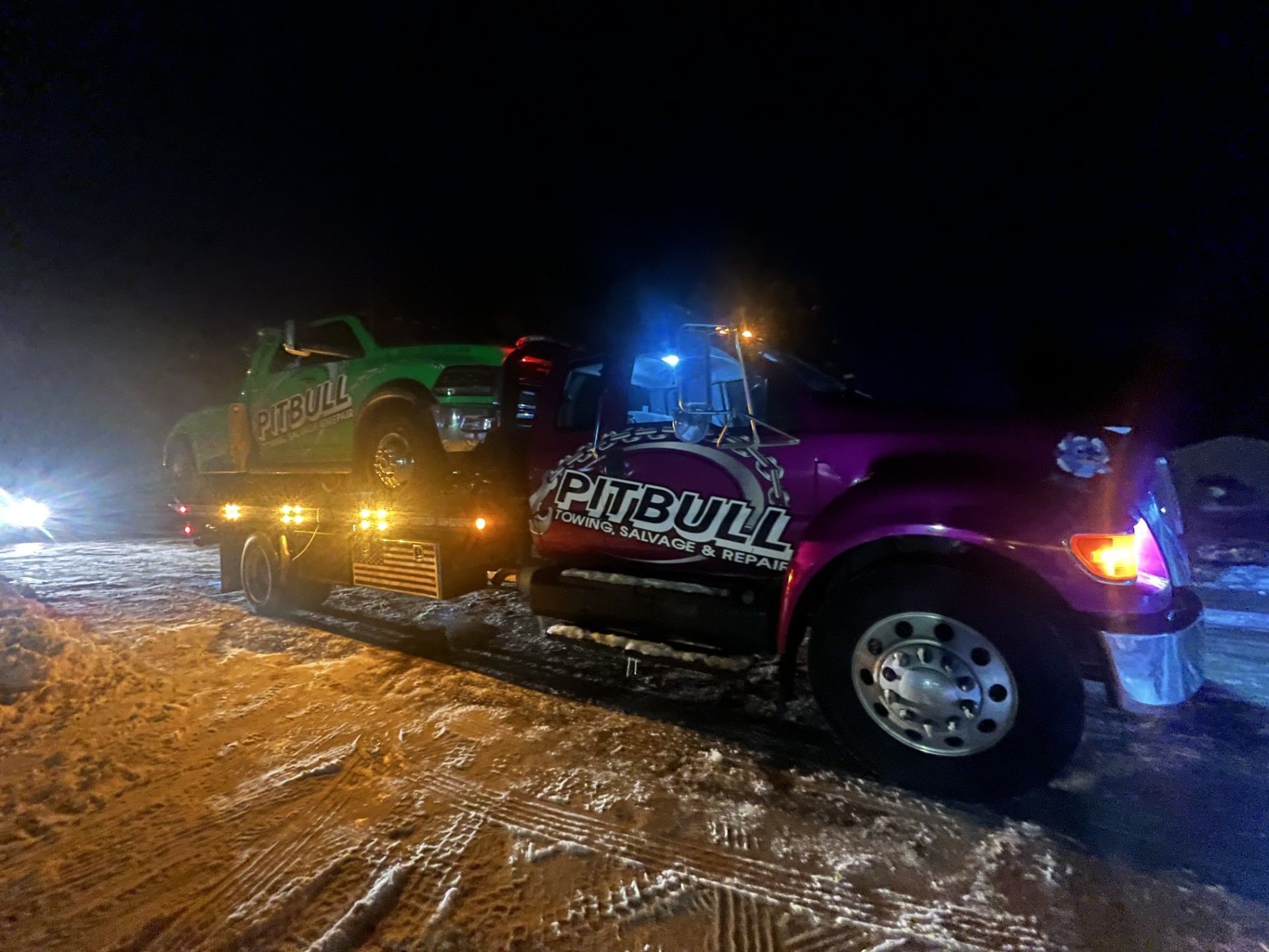 Tow truck with green car at night. Truck is purple, car is green, both have Pitbull logo.