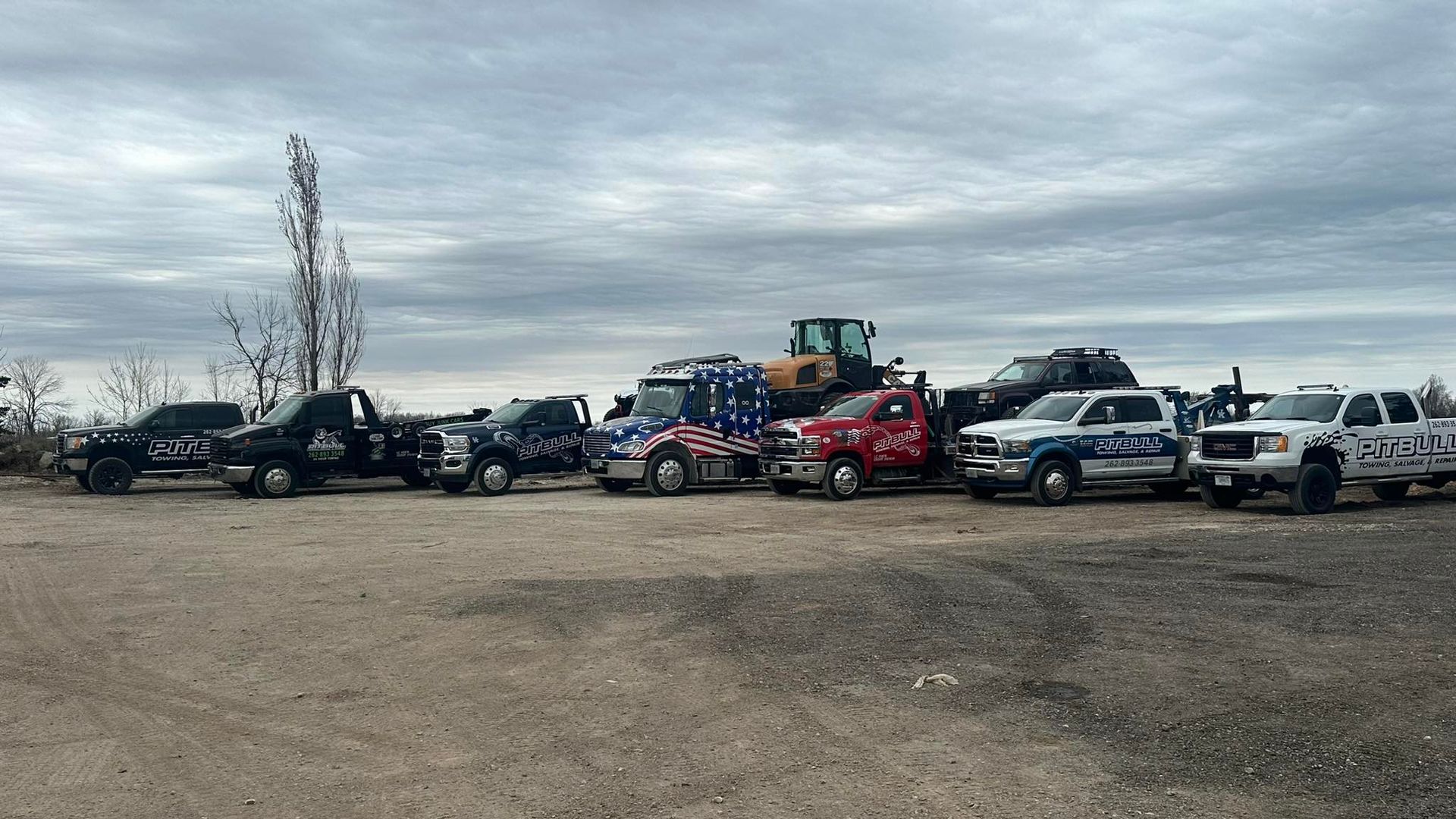 Multiple tow trucks and heavy machinery parked on gravel, cloudy sky in background.