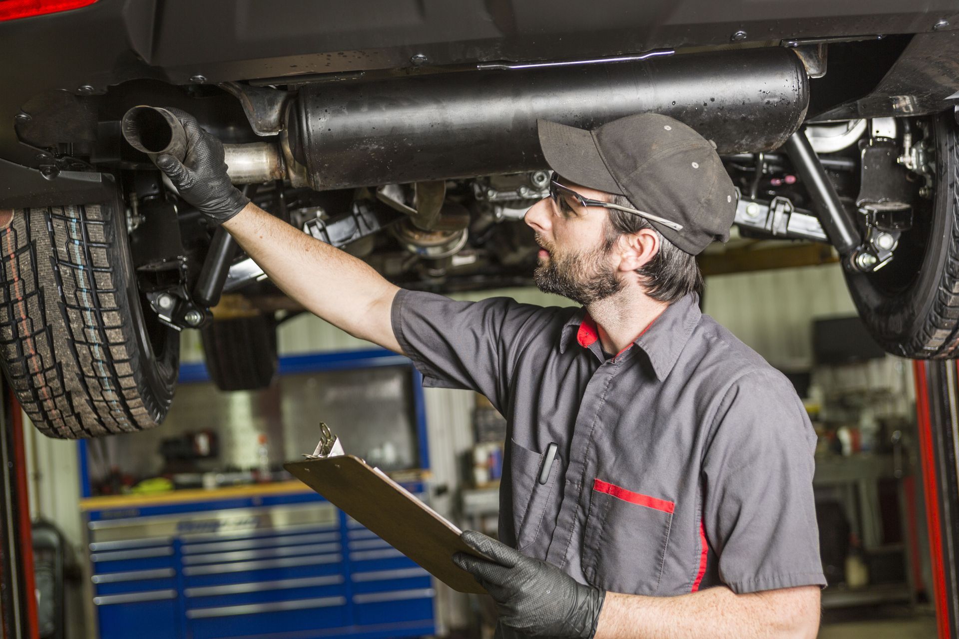 Mechanic inspecting car's undercarriage, holding clipboard. Wears gloves, cap, and glasses.
