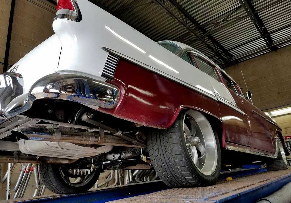 Classic two-tone red and white car on a lift, angled view from the rear. Chrome bumper and custom wheels are visible.