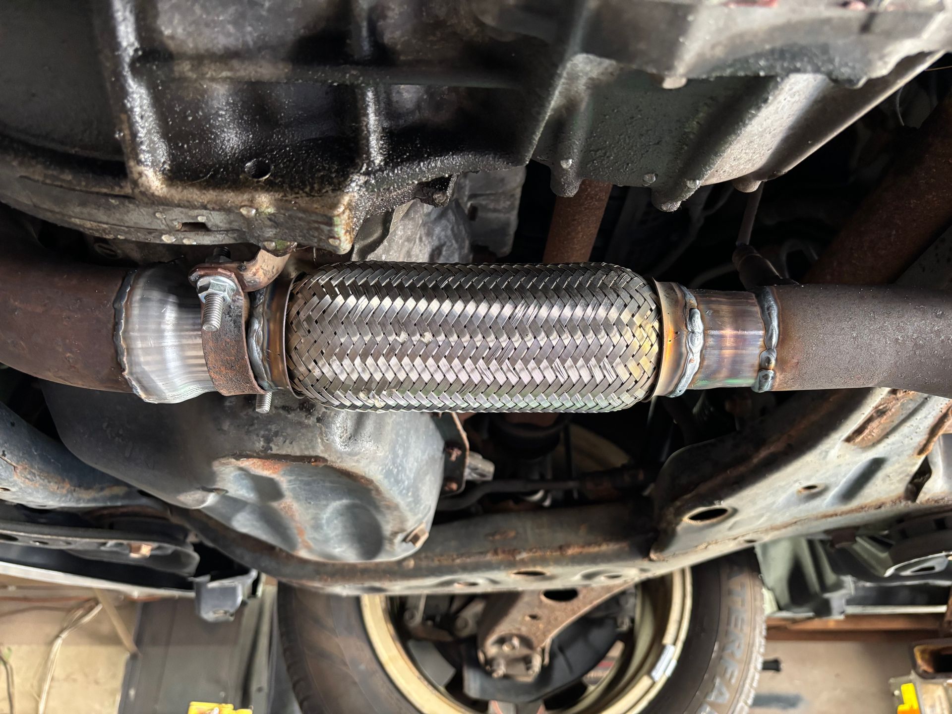 Underside view of a car's exhaust system, showing a flexible pipe section connecting two metal pipes, welded together.
